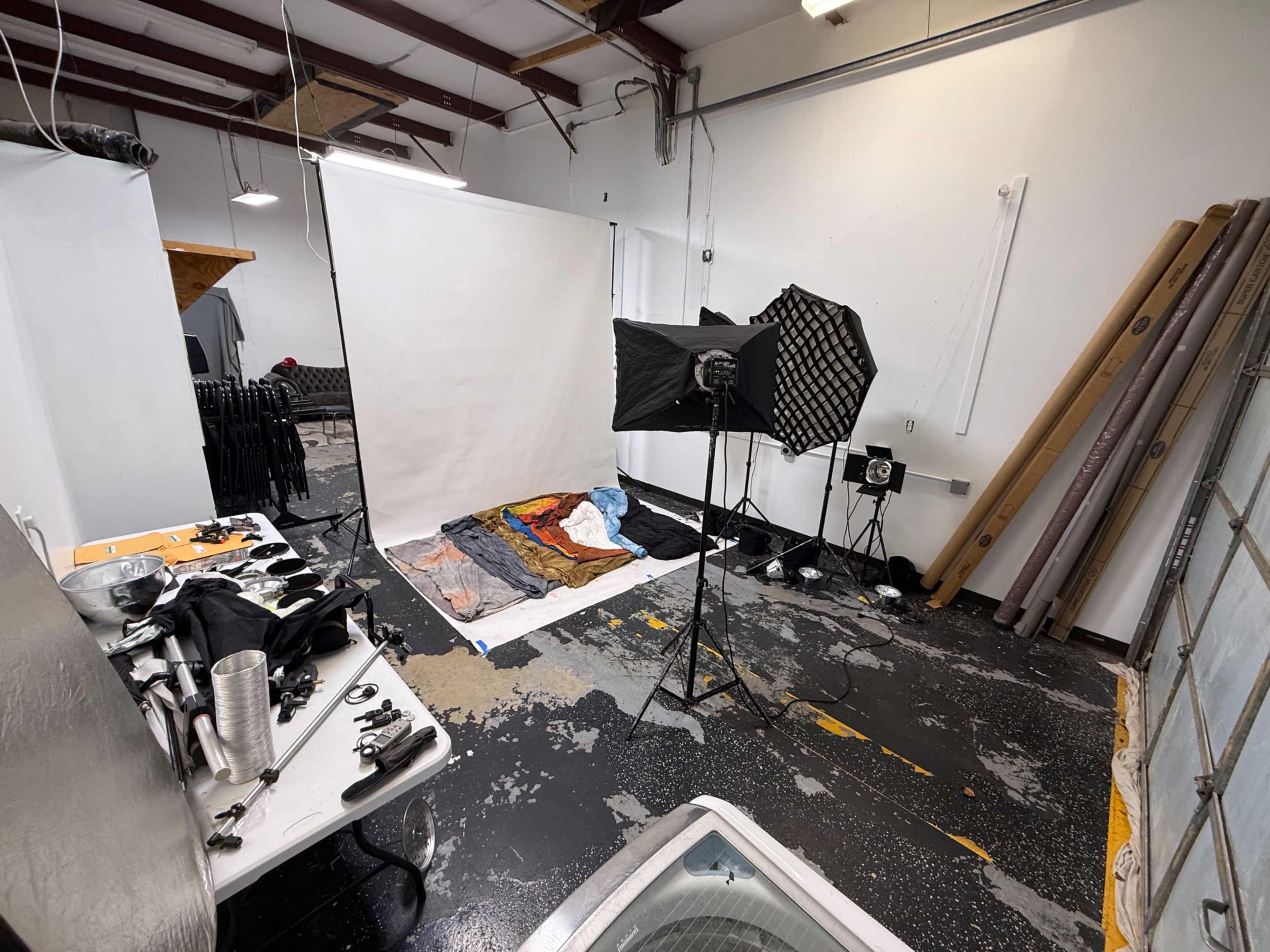 The image shows a photography studio with a backdrop, lighting equipment, and various props arranged on the floor.