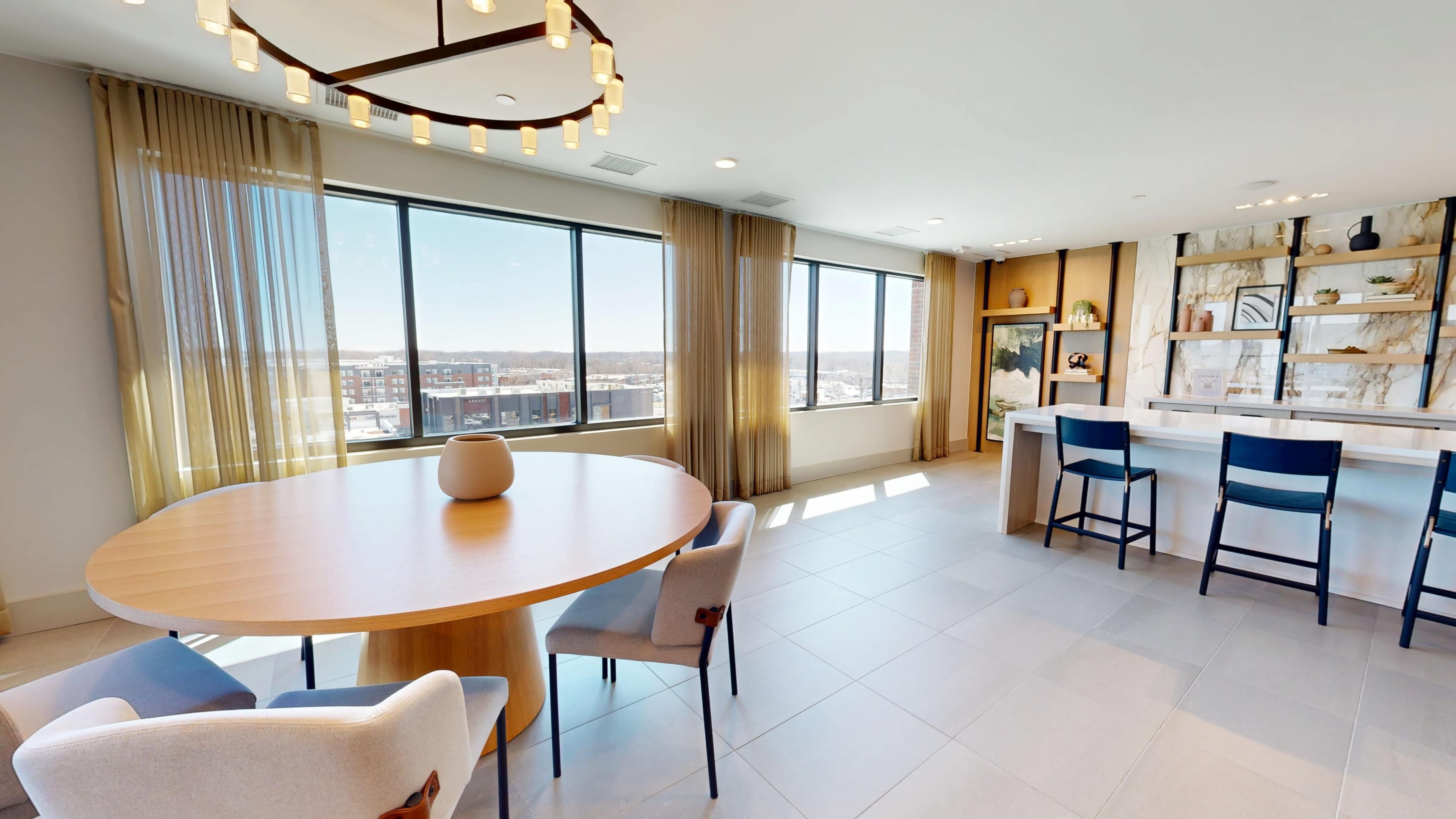 A modern dining area features a round wooden table surrounded by upholstered chairs, large windows offering a panoramic view, and a sleek kitchen in the background.