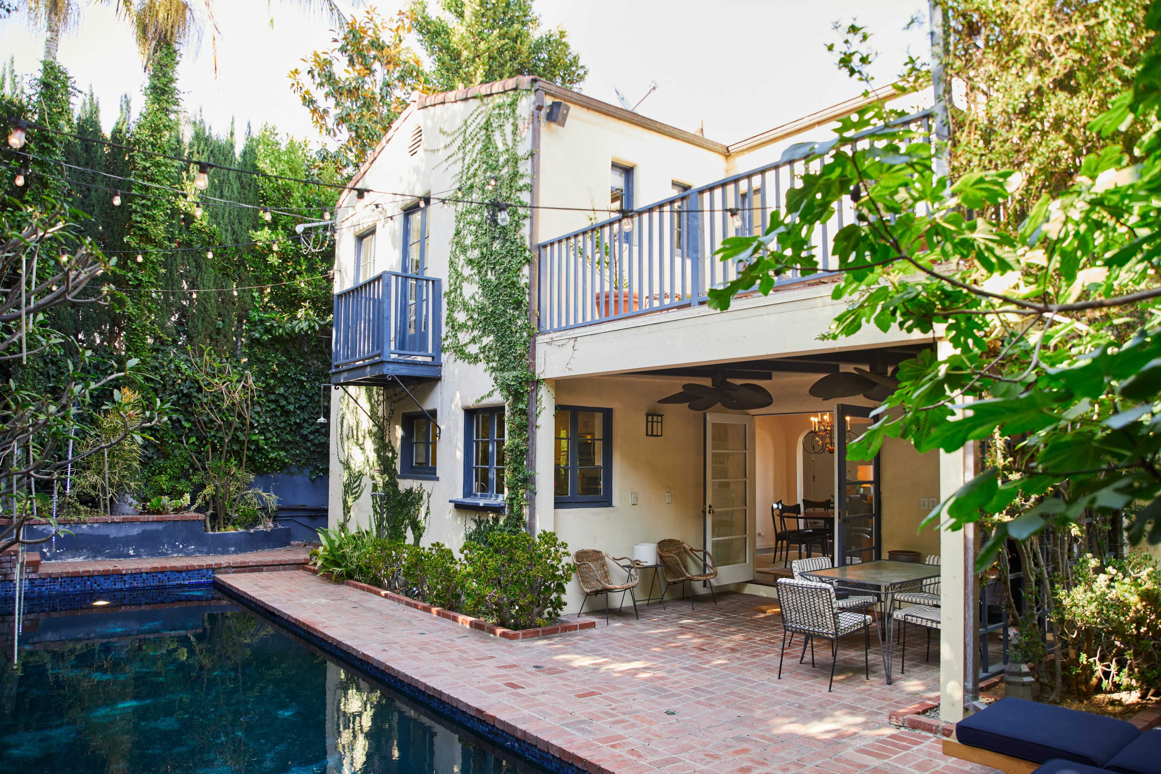 A two-story house with a pool and outdoor dining area is surrounded by greenery, featuring a balcony and patterned tile flooring.