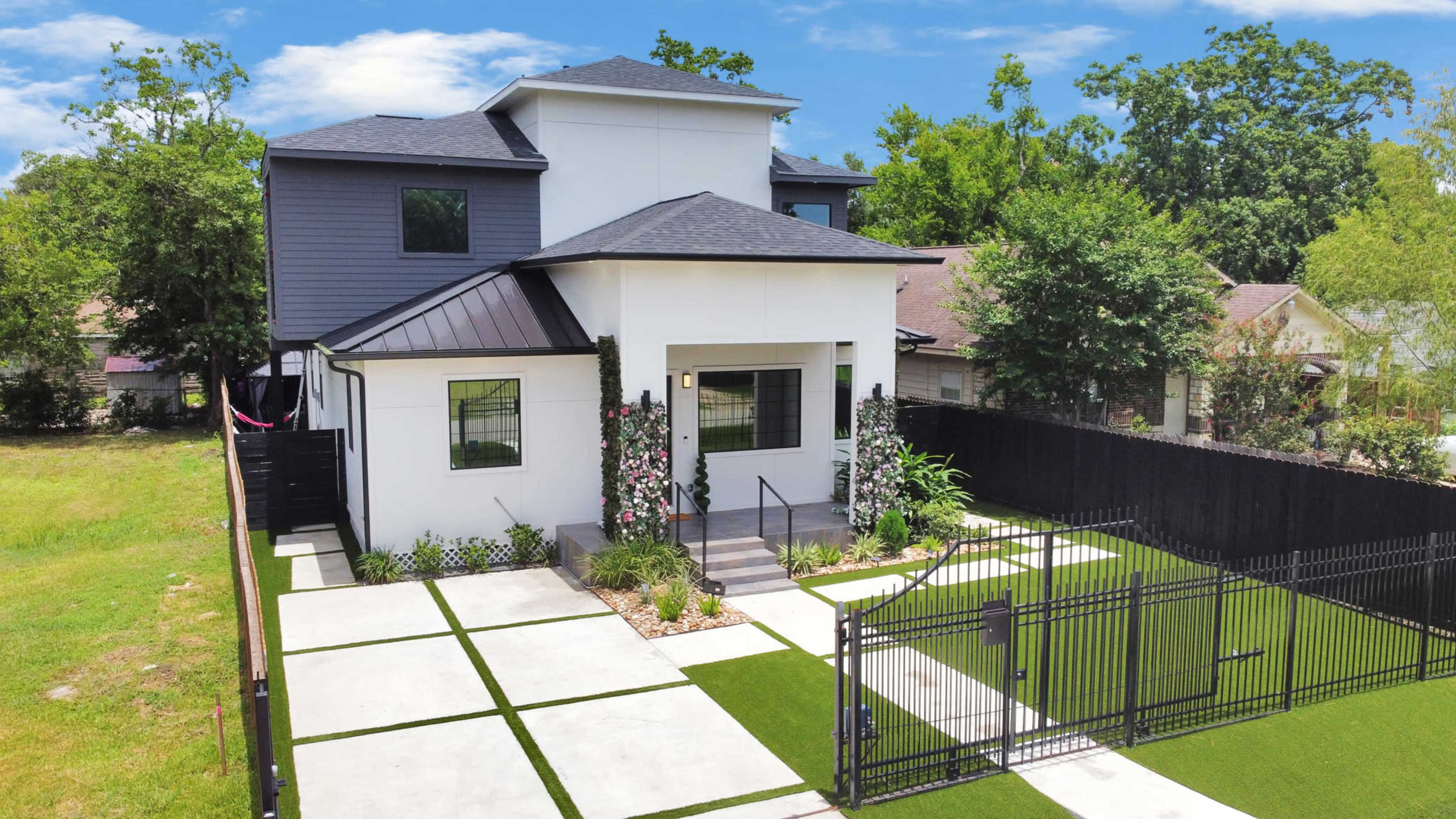 A modern two-story house with a white exterior and a gray roof is surrounded by a landscaped yard and a black fence.