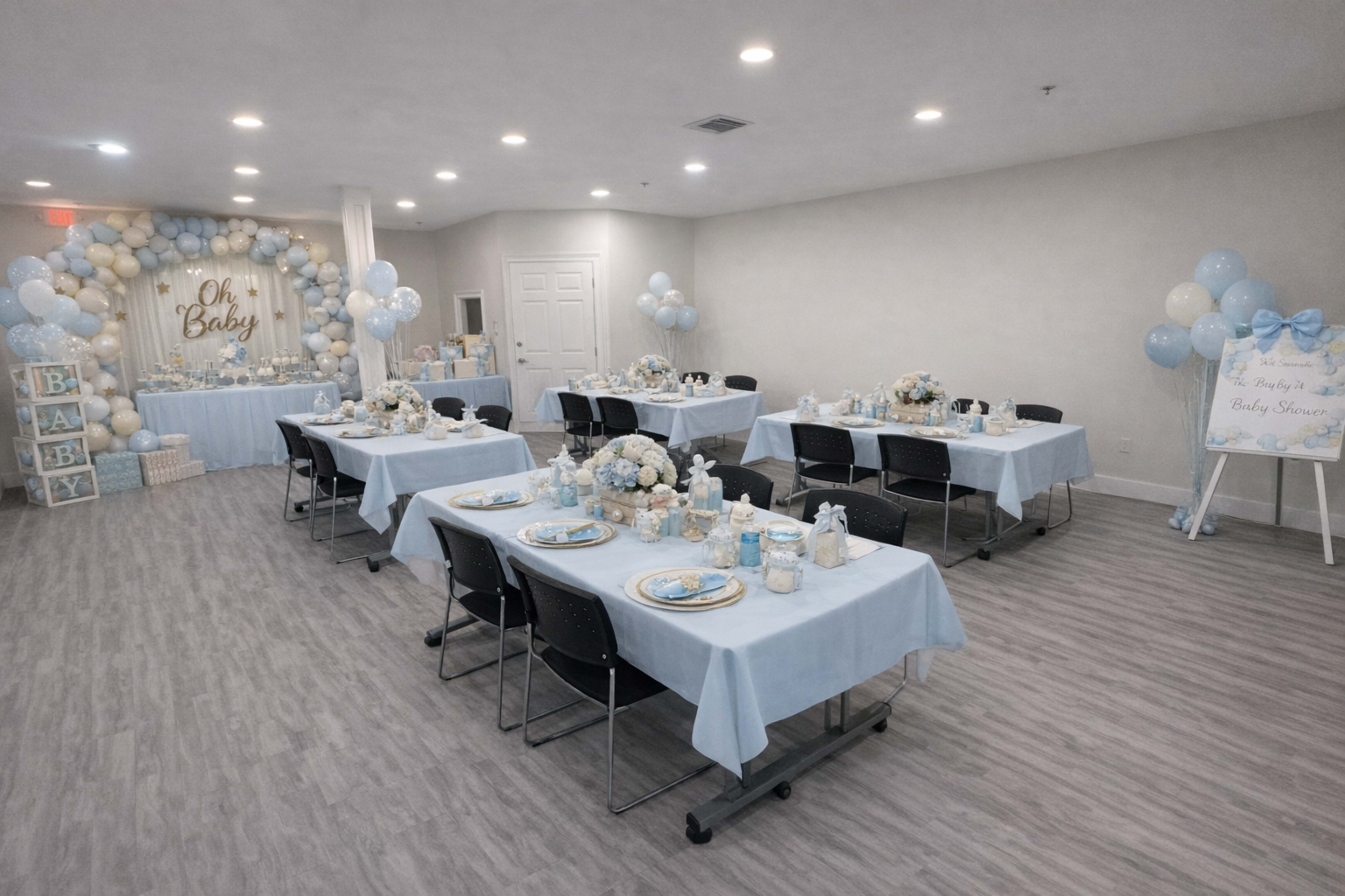 The image shows a decorated indoor space set up for a baby shower, featuring tables covered in light blue tablecloths and adorned with floral centerpieces.