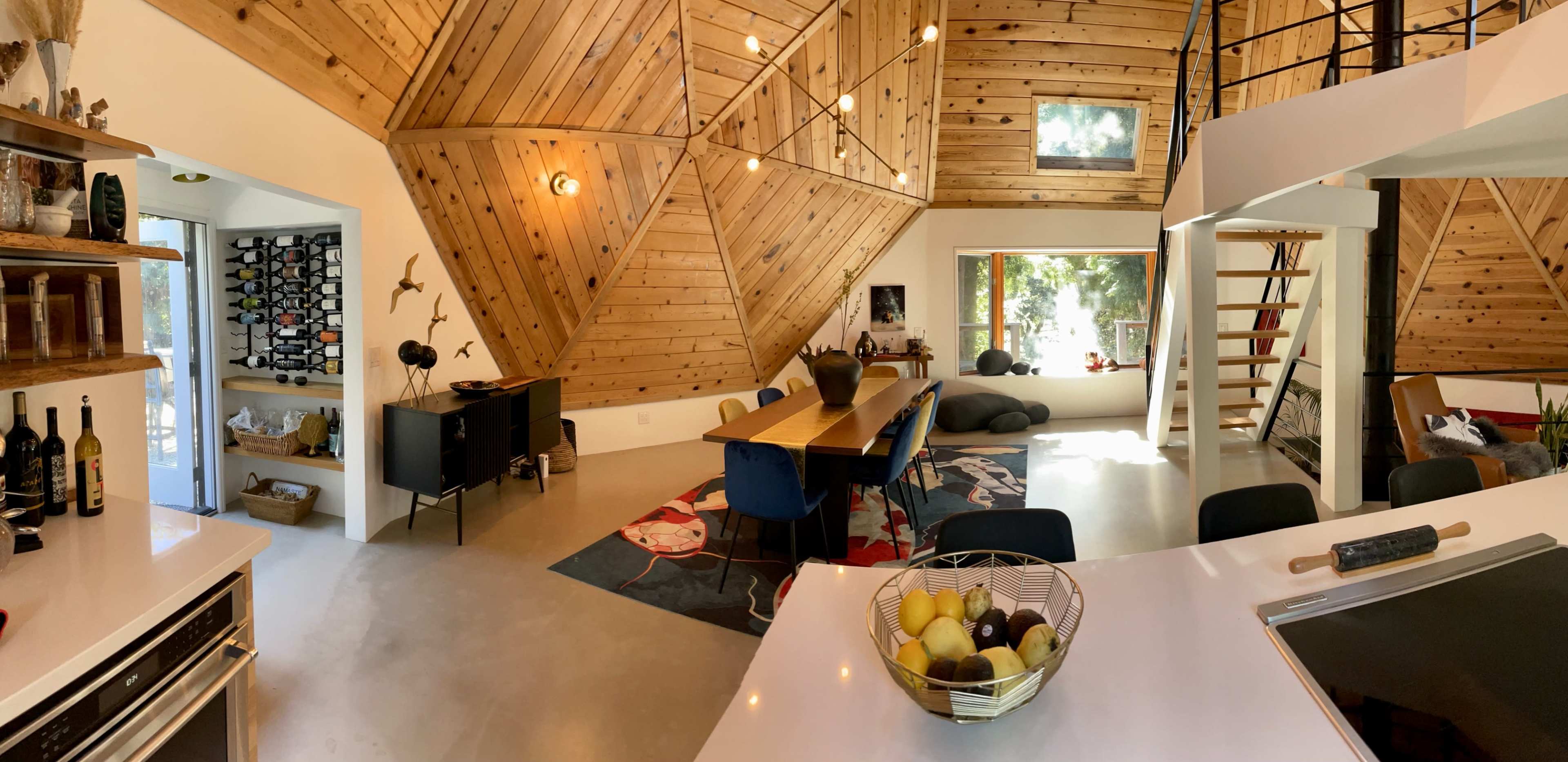 A modern, triangular-shaped interior space featuring a wooden ceiling, a long dining table with blue chairs, and a kitchen area with a basket of fruit on the counter.