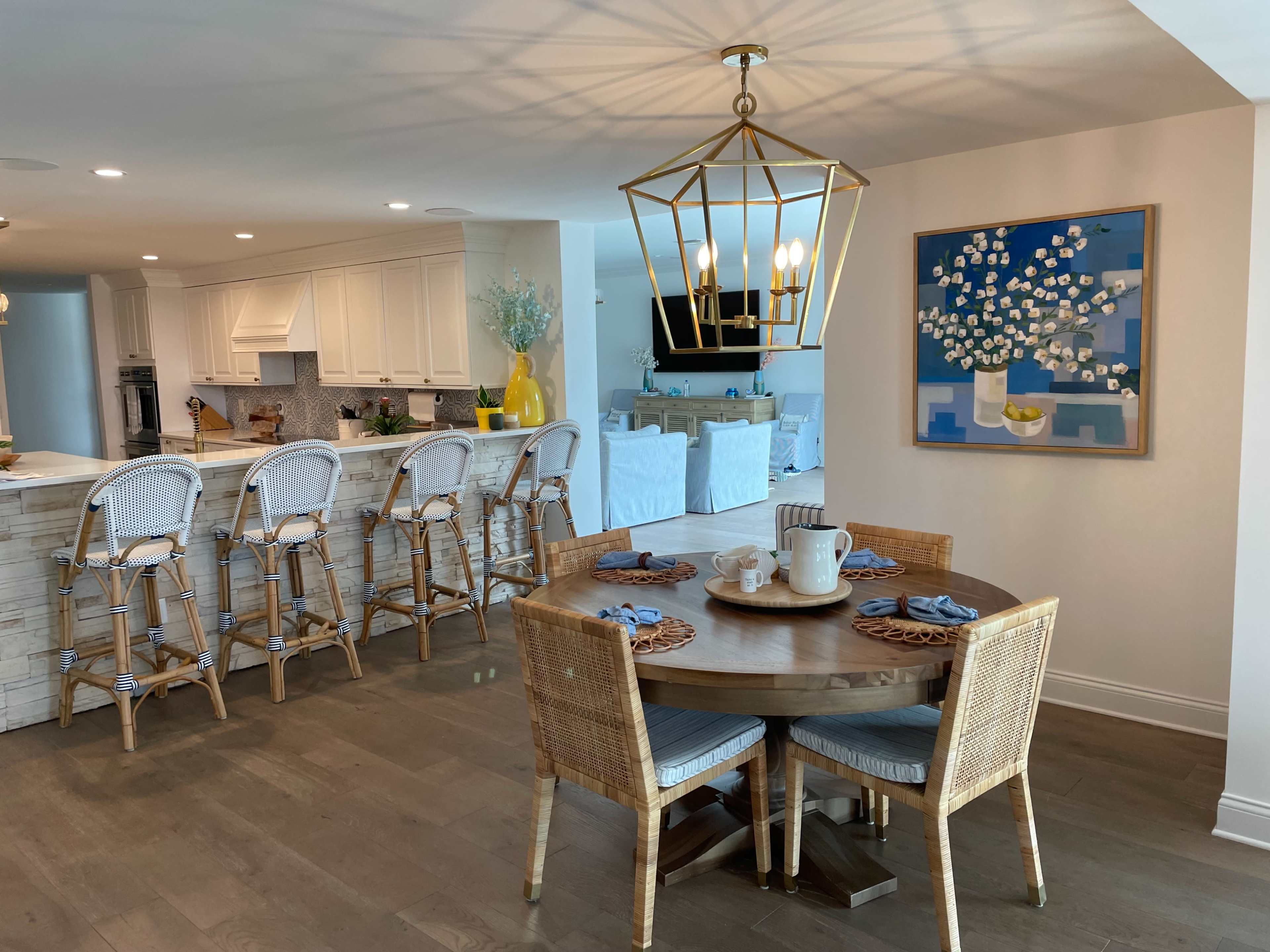 A circular wooden dining table with four wicker chairs is set with dishware, facing a kitchen bar with high stools and a decorative painting on the wall.