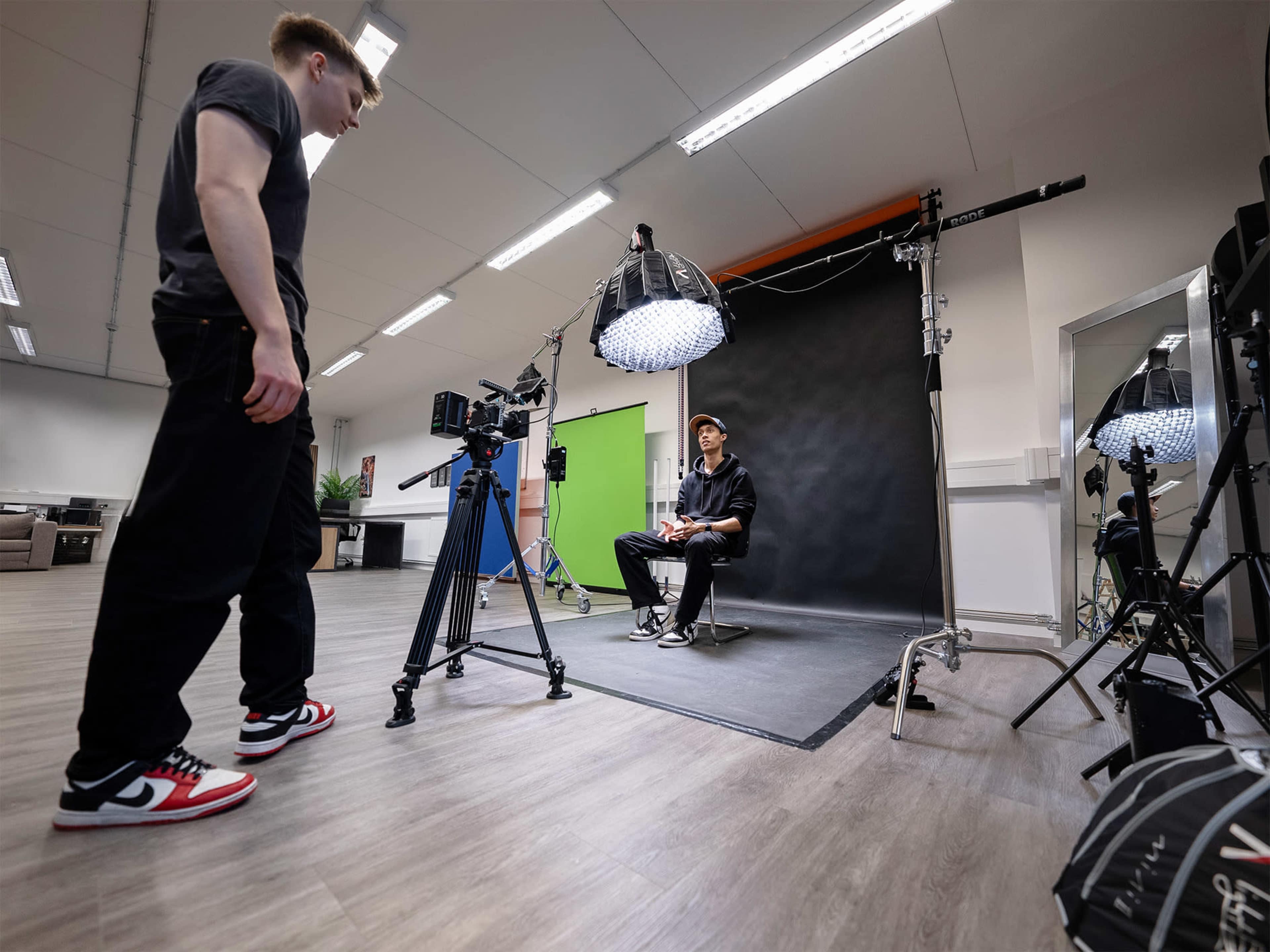 A person sits on a chair in front of a large lighting setup while another person adjusts a camera in a studio.