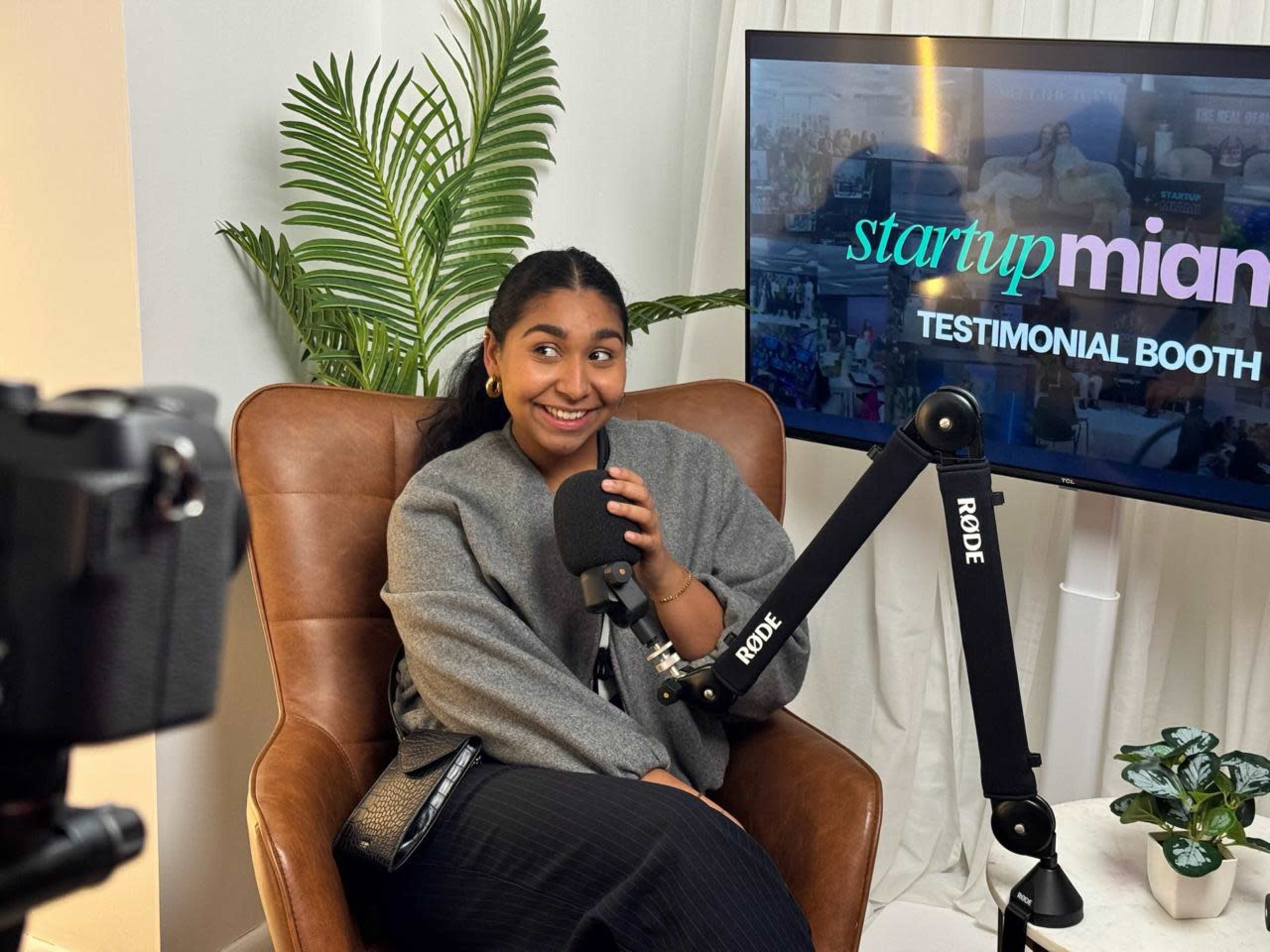A woman sits in a leather chair holding a microphone in a studio setting with a screen displaying "startup miami testimonial booth" in the background.