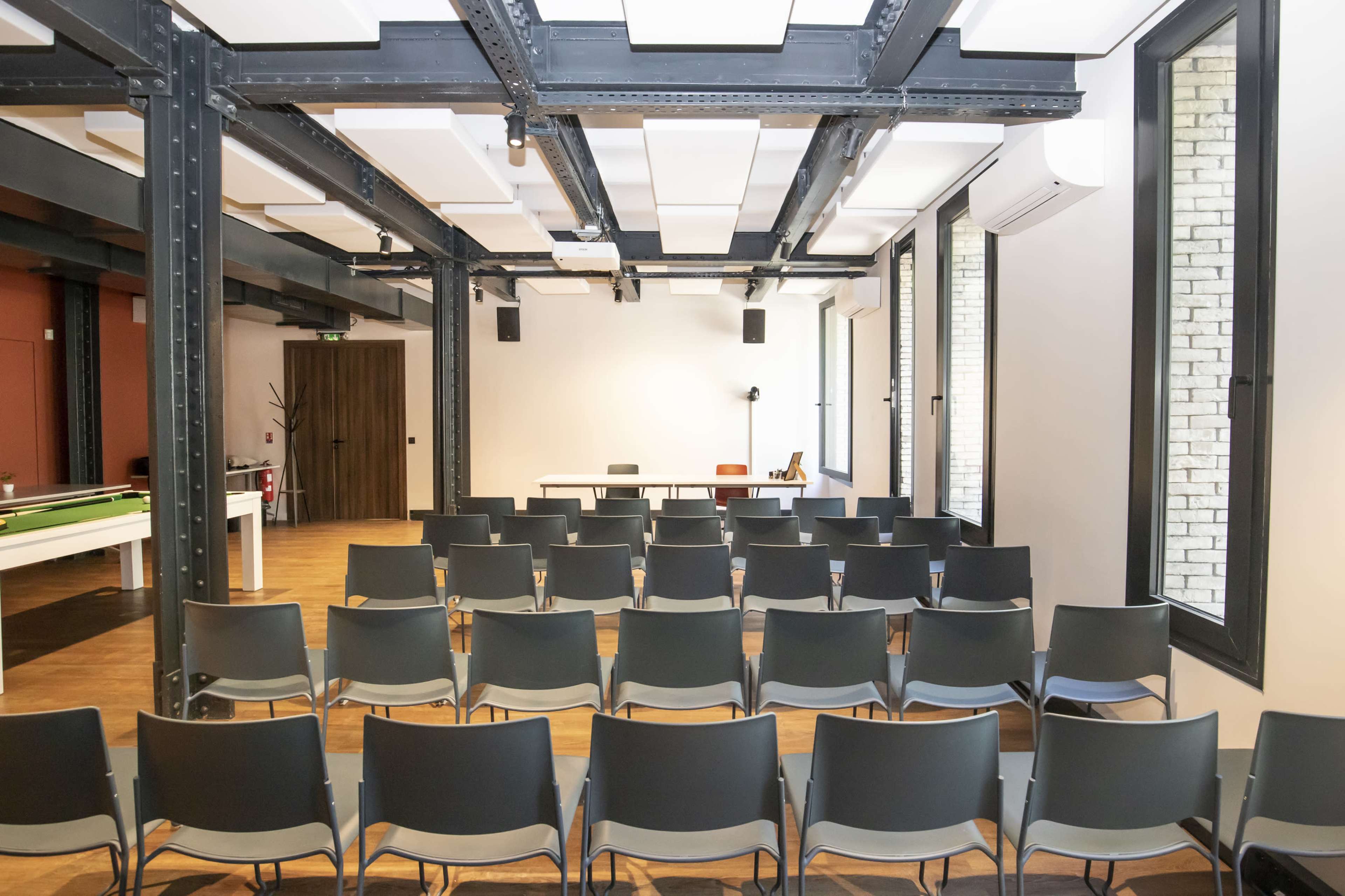 The image shows a conference room set up with rows of gray chairs facing a blank screen and a table at the front.