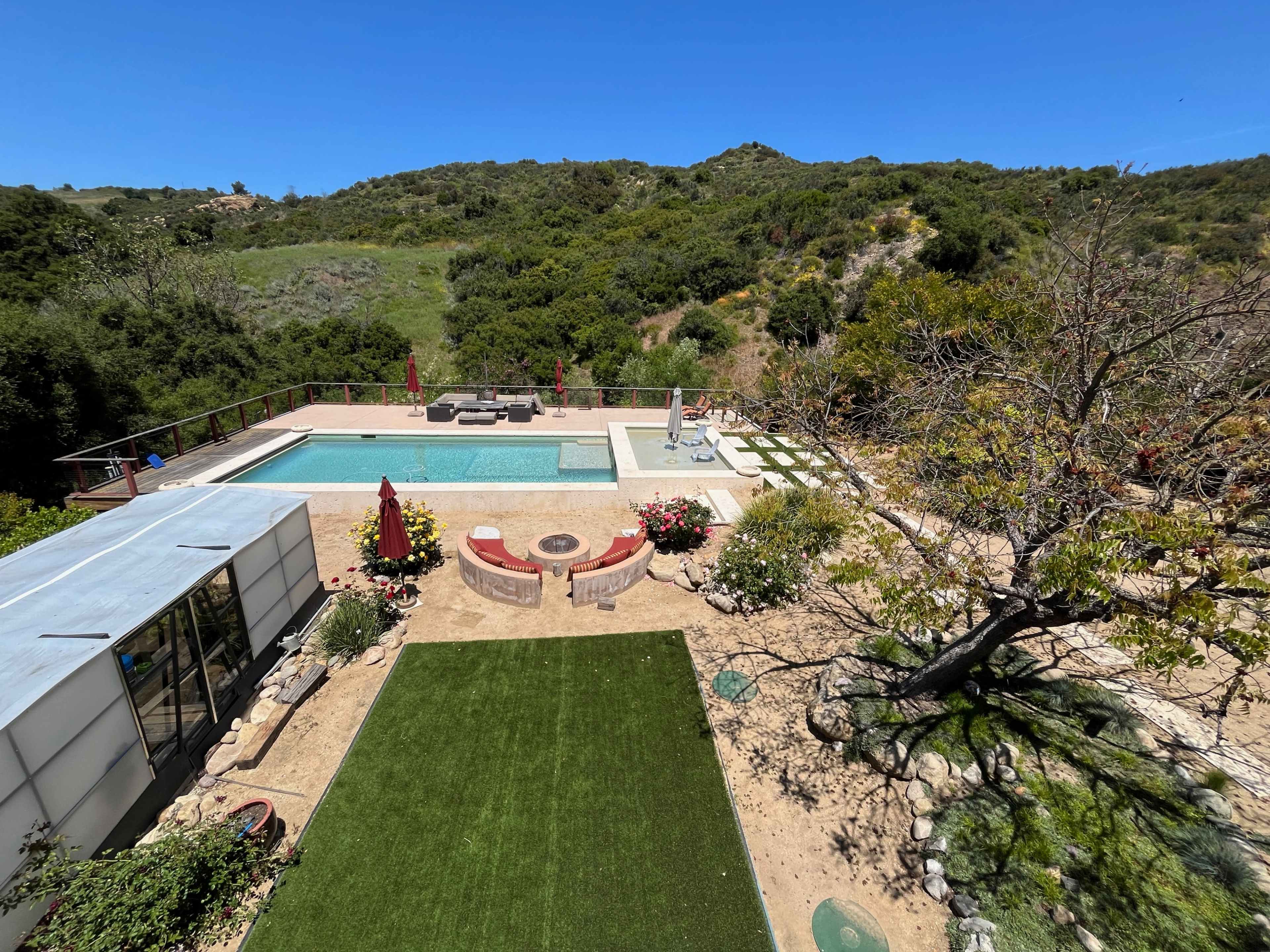 A modern backyard features a swimming pool surrounded by lounge furniture, a landscaped area with grass and flower beds, and views of a hillside.