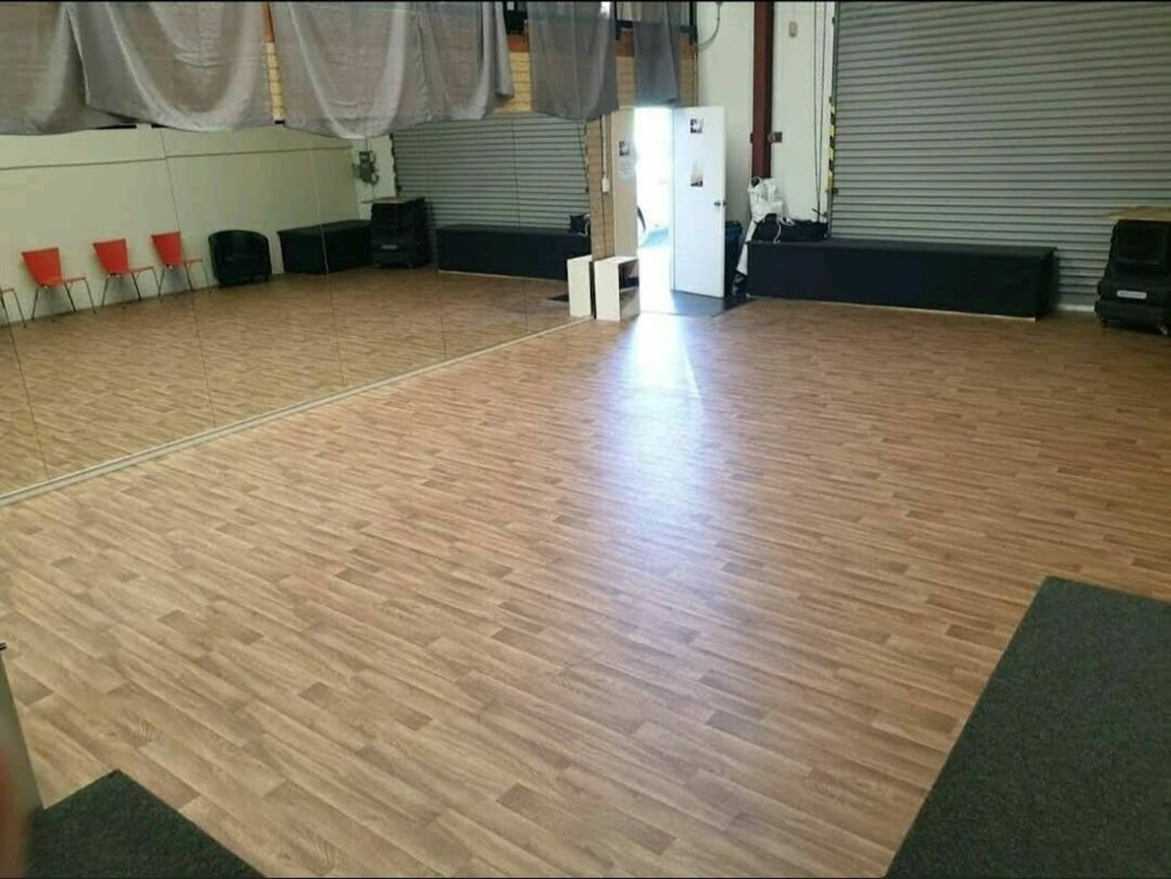 A spacious dance studio with wooden flooring, mirrors on one wall, and red chairs along the side.