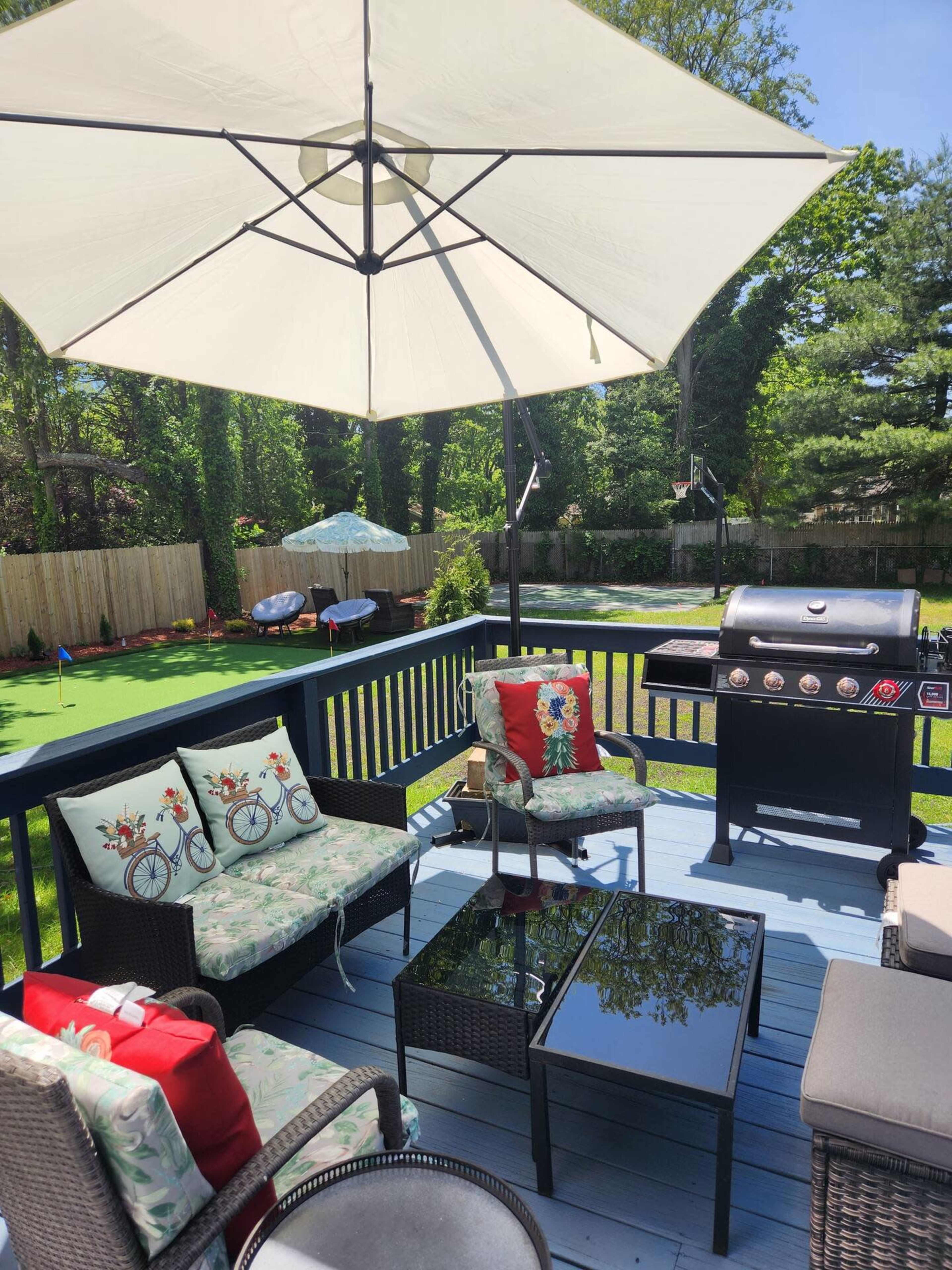 A backyard deck area with a grill, outdoor seating, and a large umbrella, surrounded by a lawn and trees.