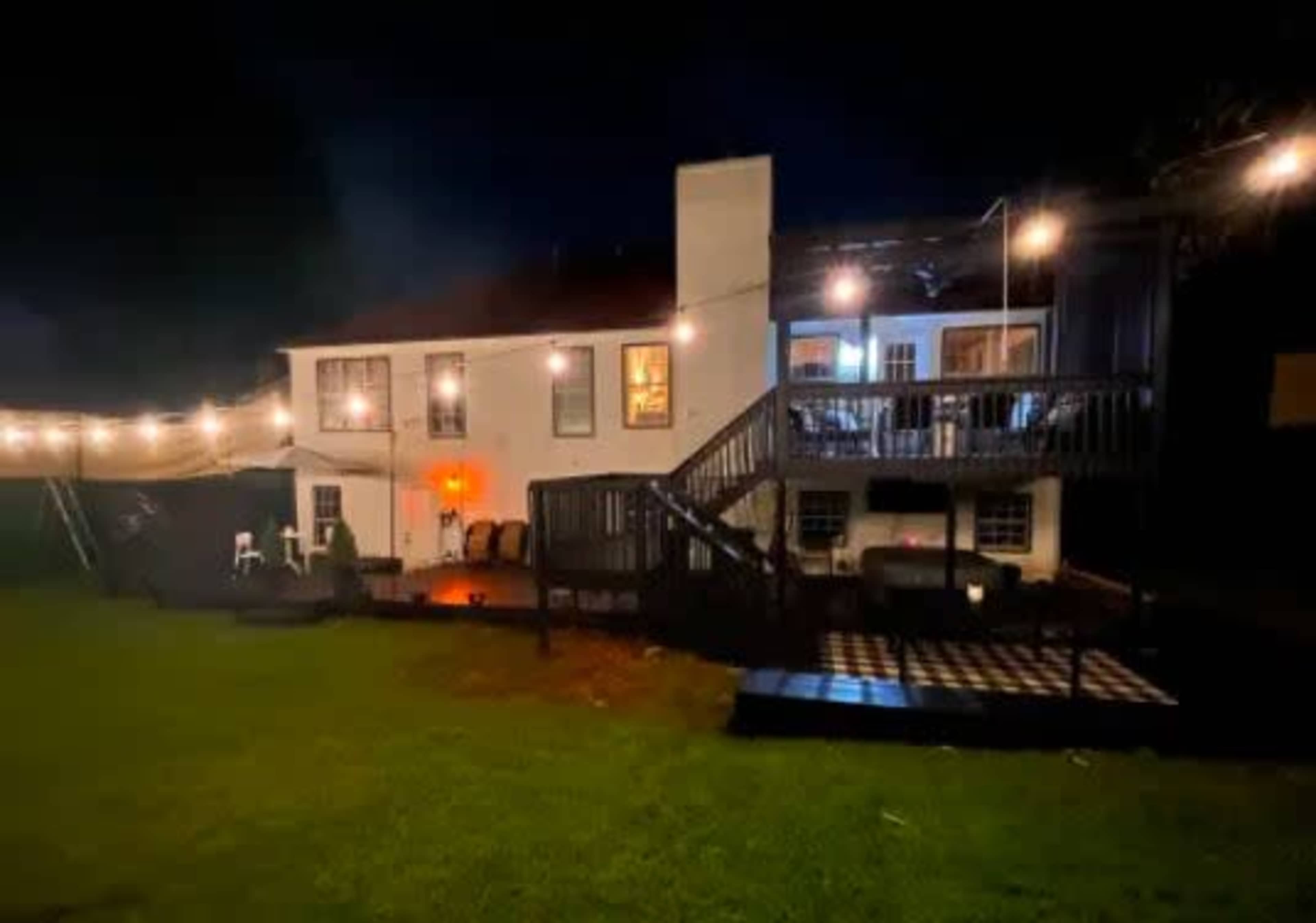 A two-story house with a deck and string lights is illuminated at night in a grassy backyard.