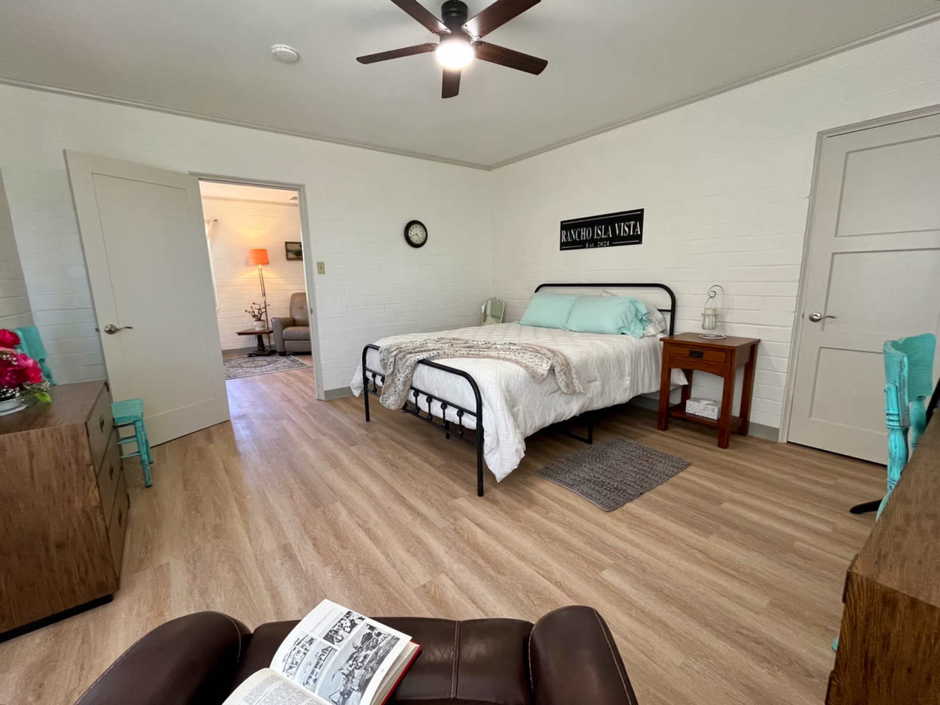 The image shows a well-furnished bedroom featuring a bed with a light-colored duvet, a bedside table, a wooden dresser, and a chair, with a view into a connected living area.