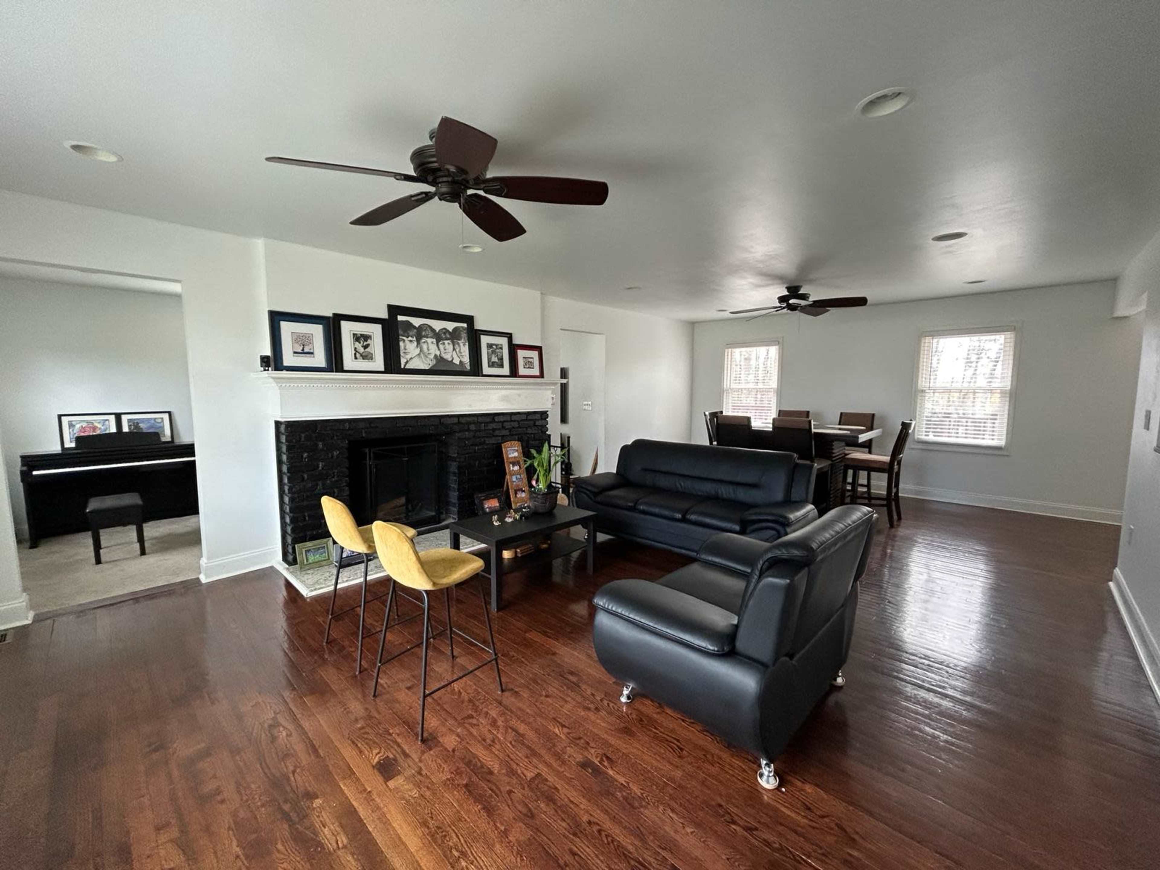 The living room features a black leather sofa, a coffee table, and a dining area, with a piano visible in the background and two ceiling fans overhead.