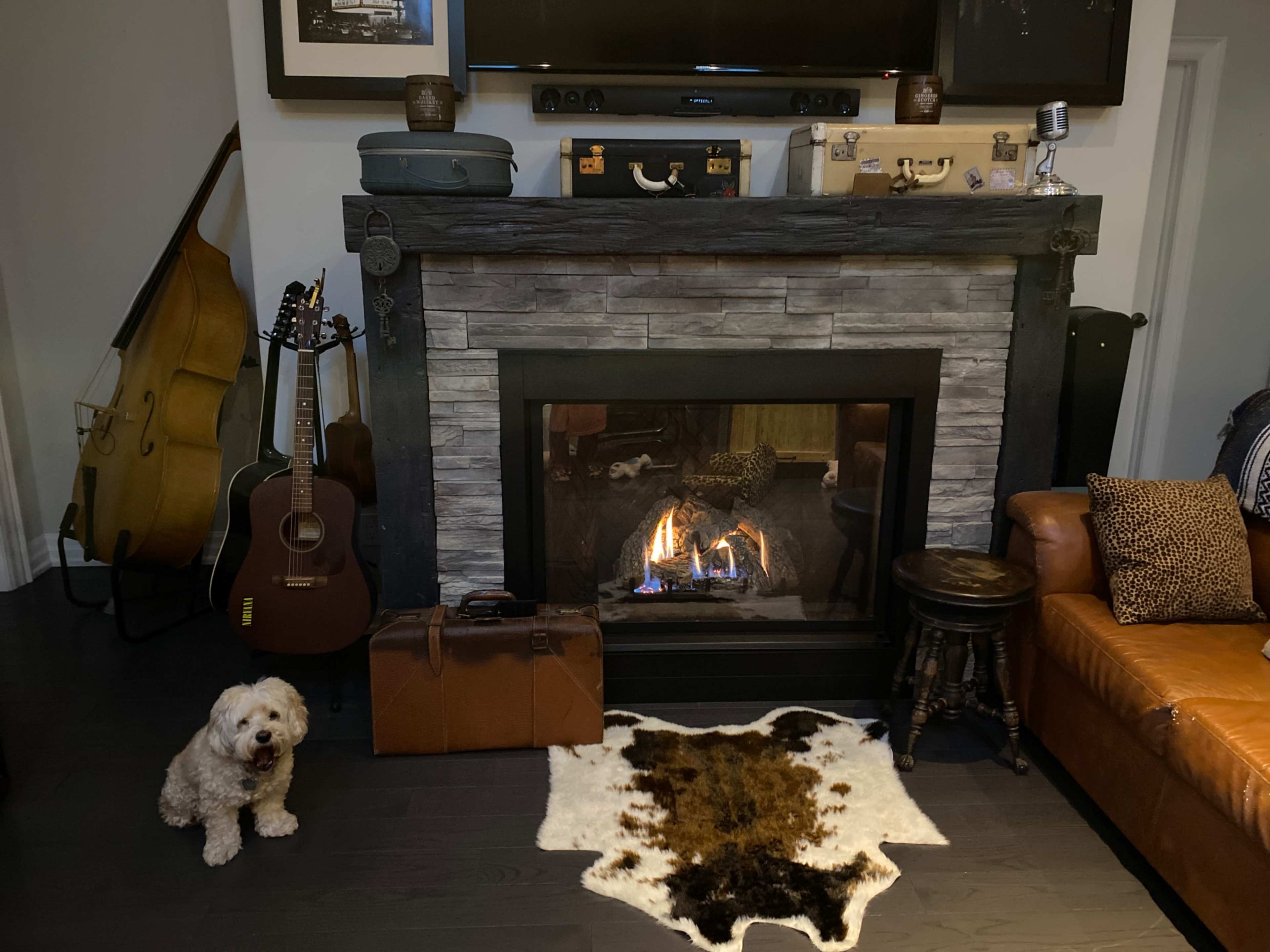 A cozy living room features a stone fireplace with a fire burning, surrounded by a brown sofa, musical instruments, and a decorative cowhide rug.