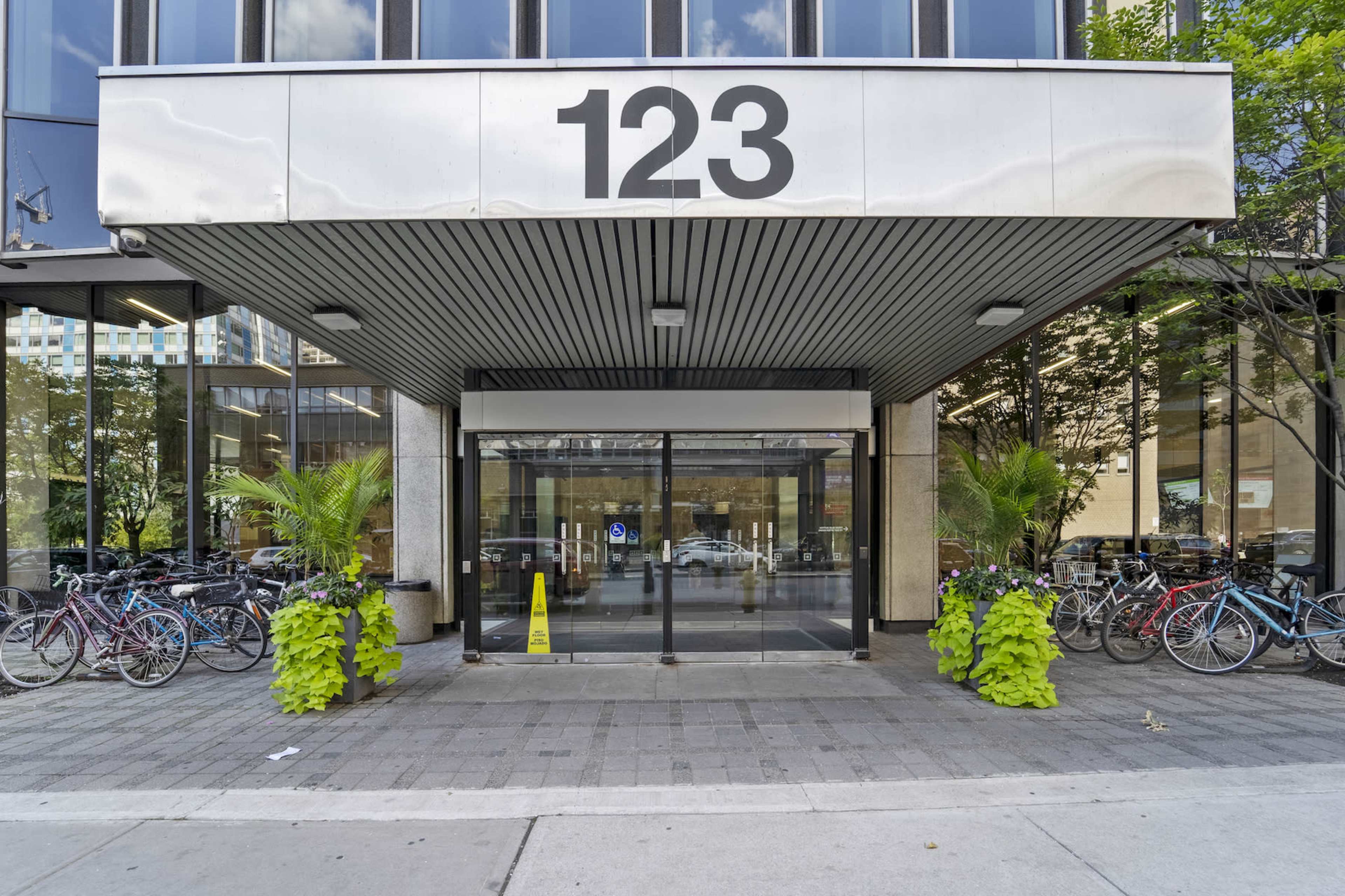 The entrance of a building at 123, with glass doors and bicycles parked on either side, surrounded by greenery.