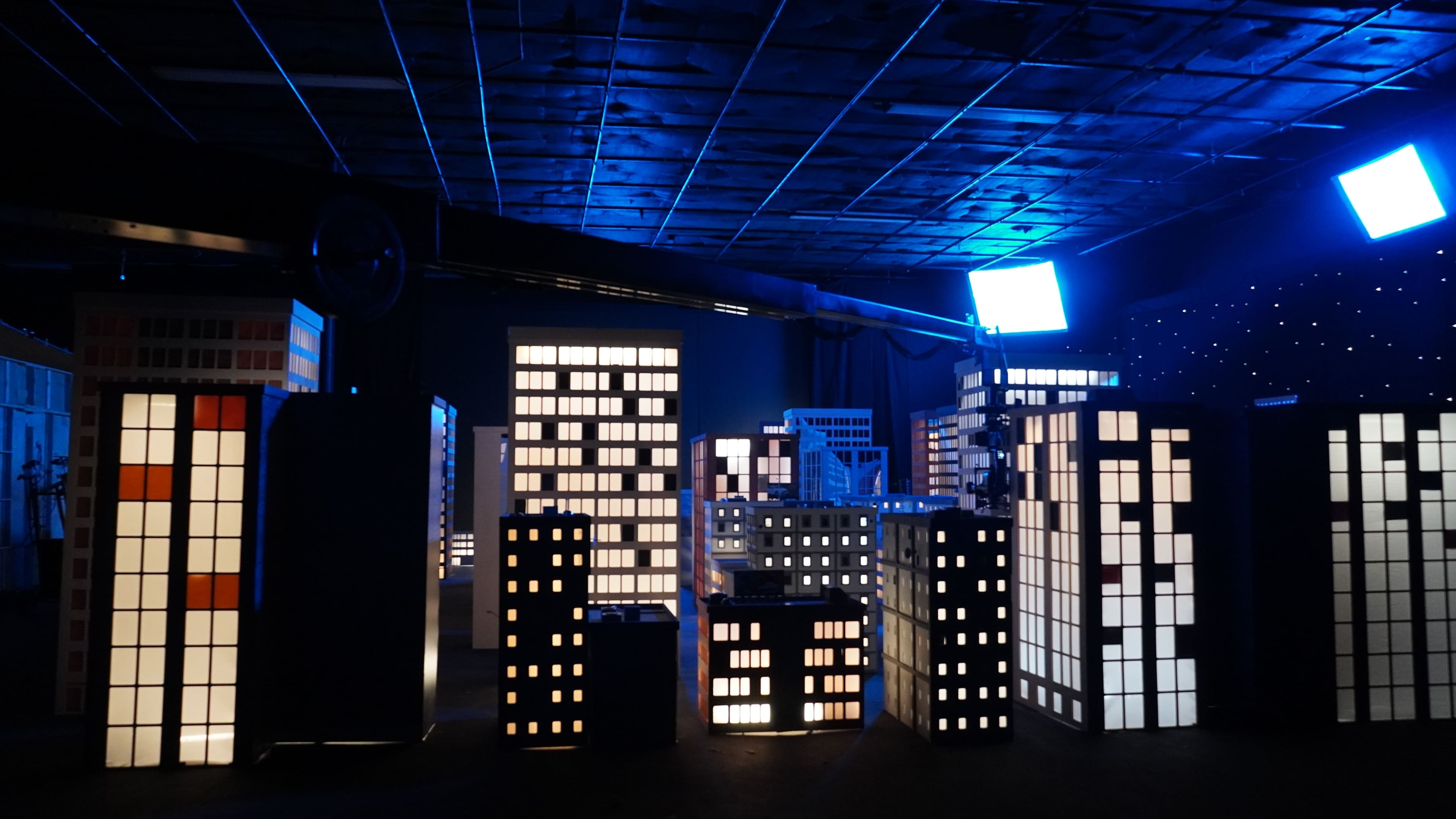 The scene features an art installation of illuminated building models arranged in a dark room.