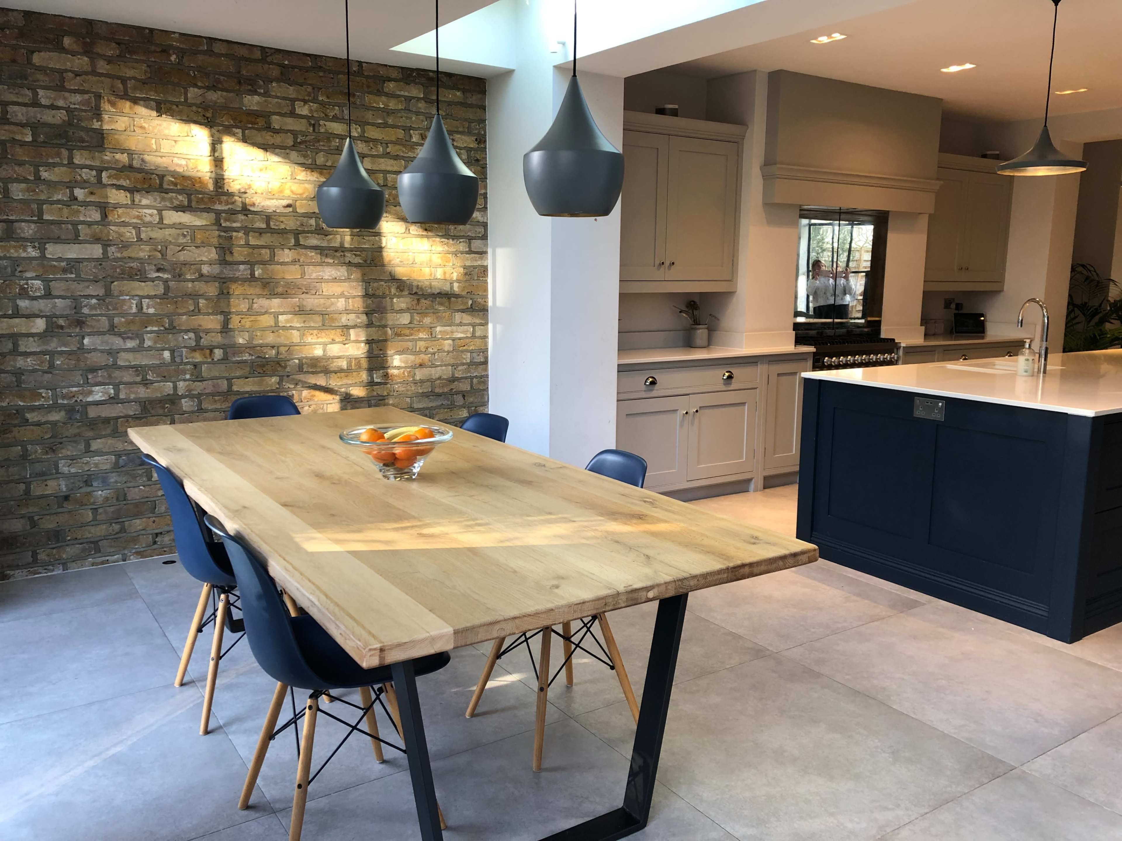 The image shows a modern kitchen and dining area featuring a large wooden table surrounded by chairs, with a brick wall in the background and pendant lighting overhead.