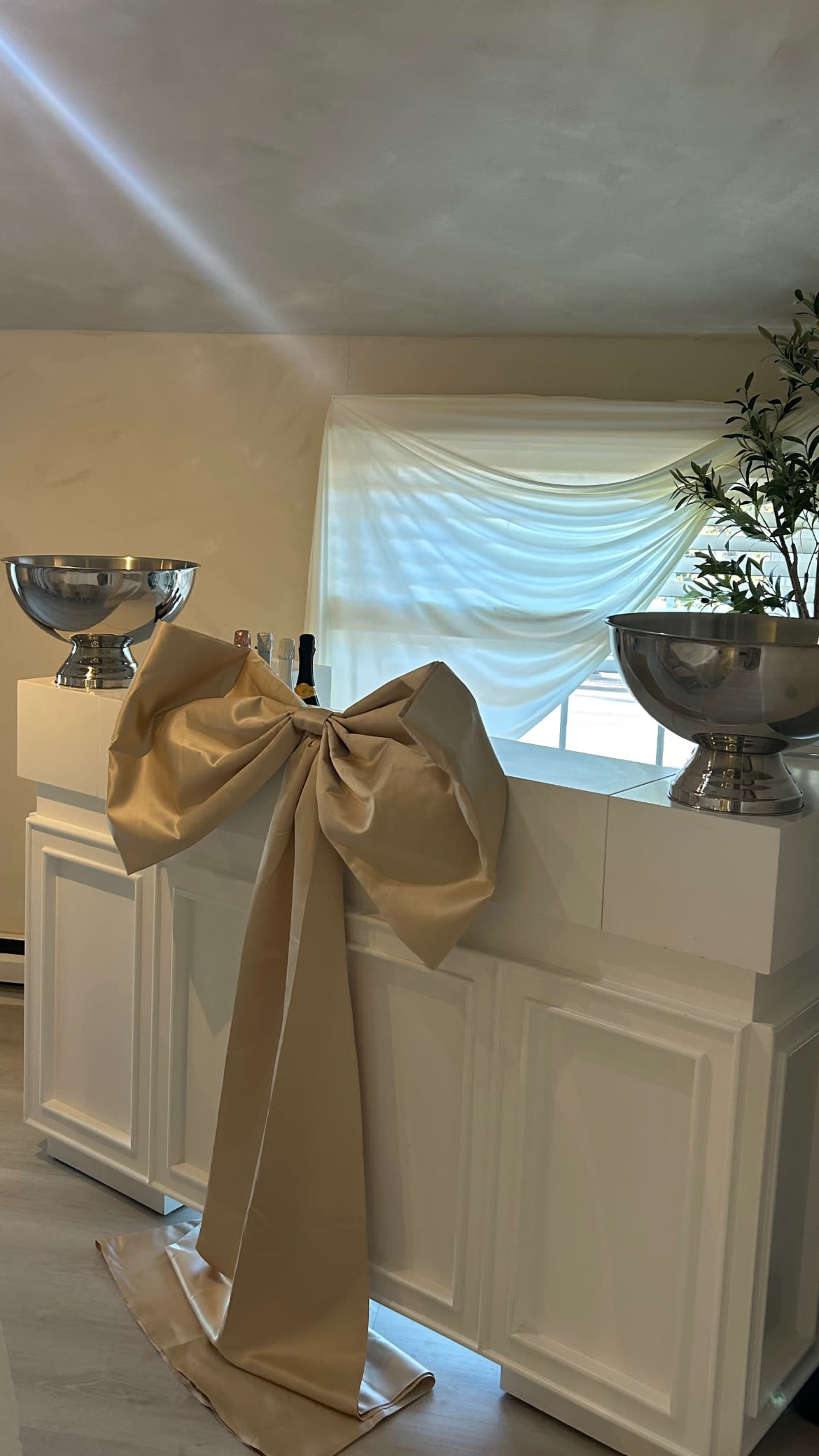The image features a bar setup with two metal bowls on either side of a decorative bow made of beige fabric, against a backdrop of a window with draped curtains.
