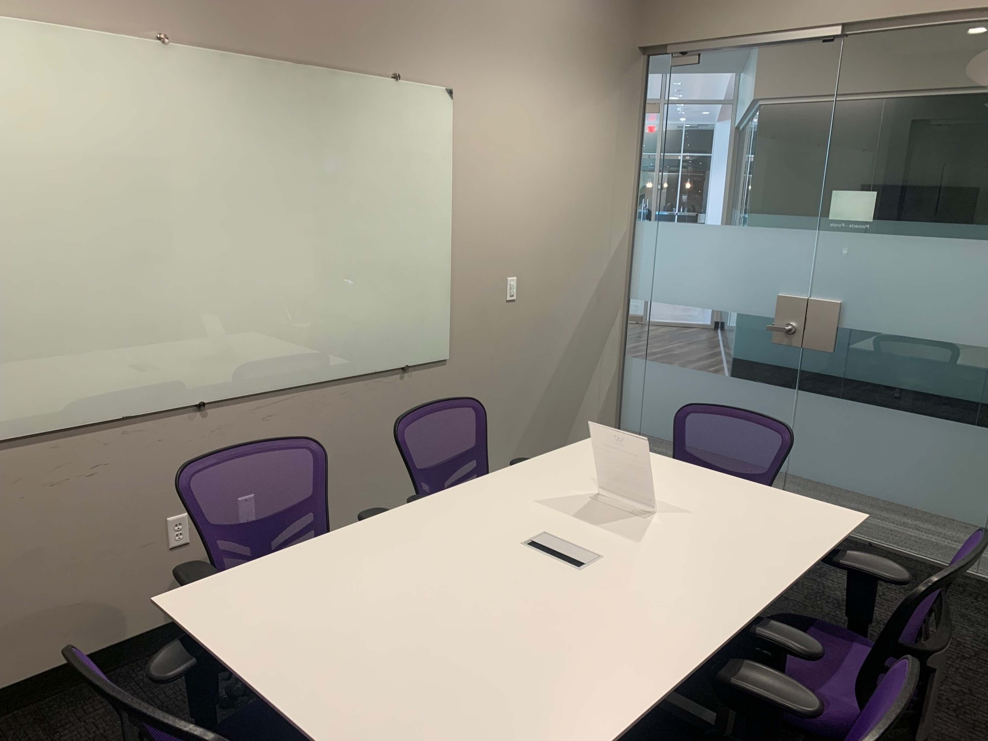A conference room features a whiteboard, a rectangular table with a central device, and four purple chairs around it.