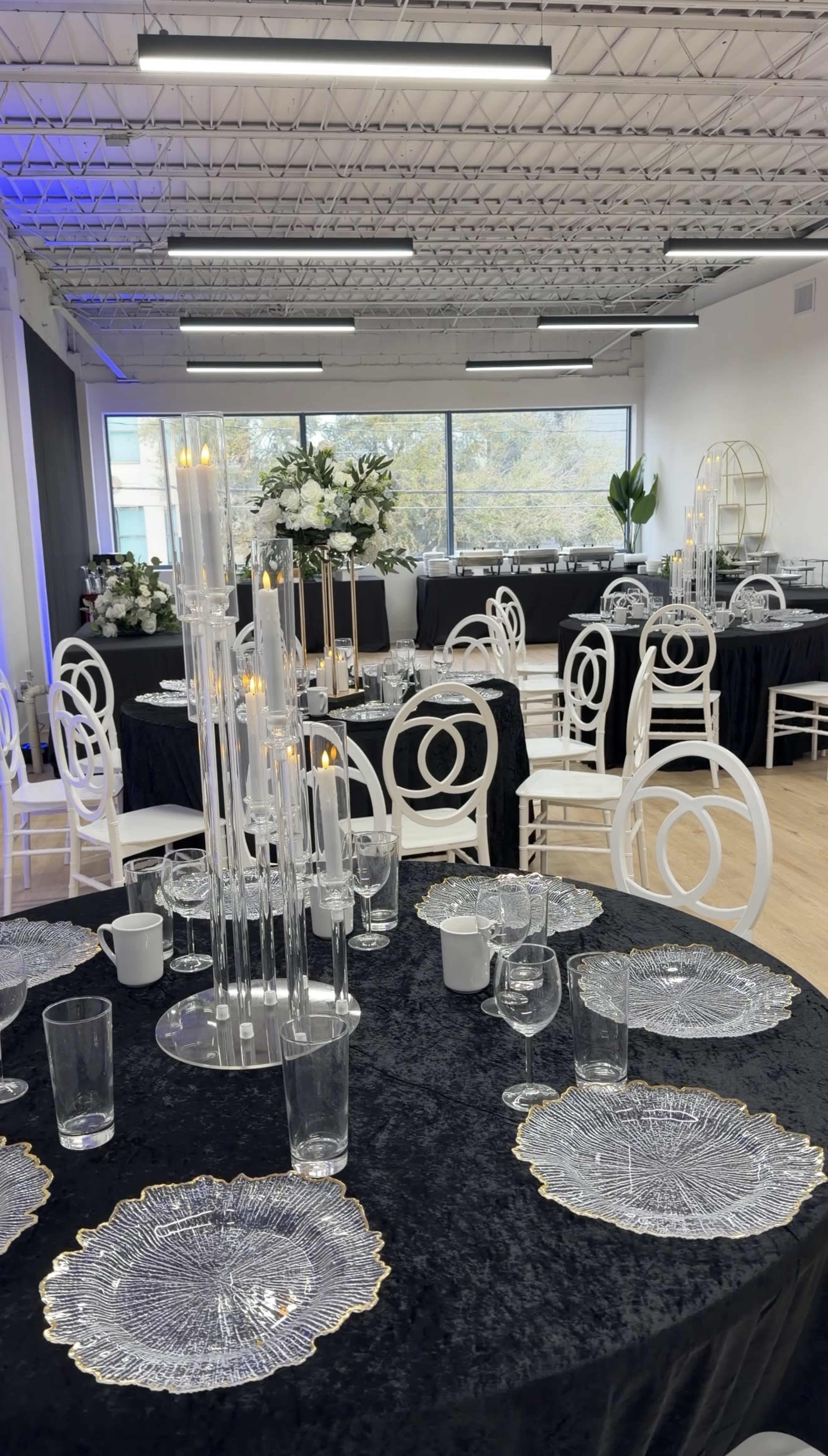 The image shows a decorated event space featuring elegantly set tables with black tablecloths, clear glassware, and candle centerpieces, alongside floral arrangements and banquet seating.
