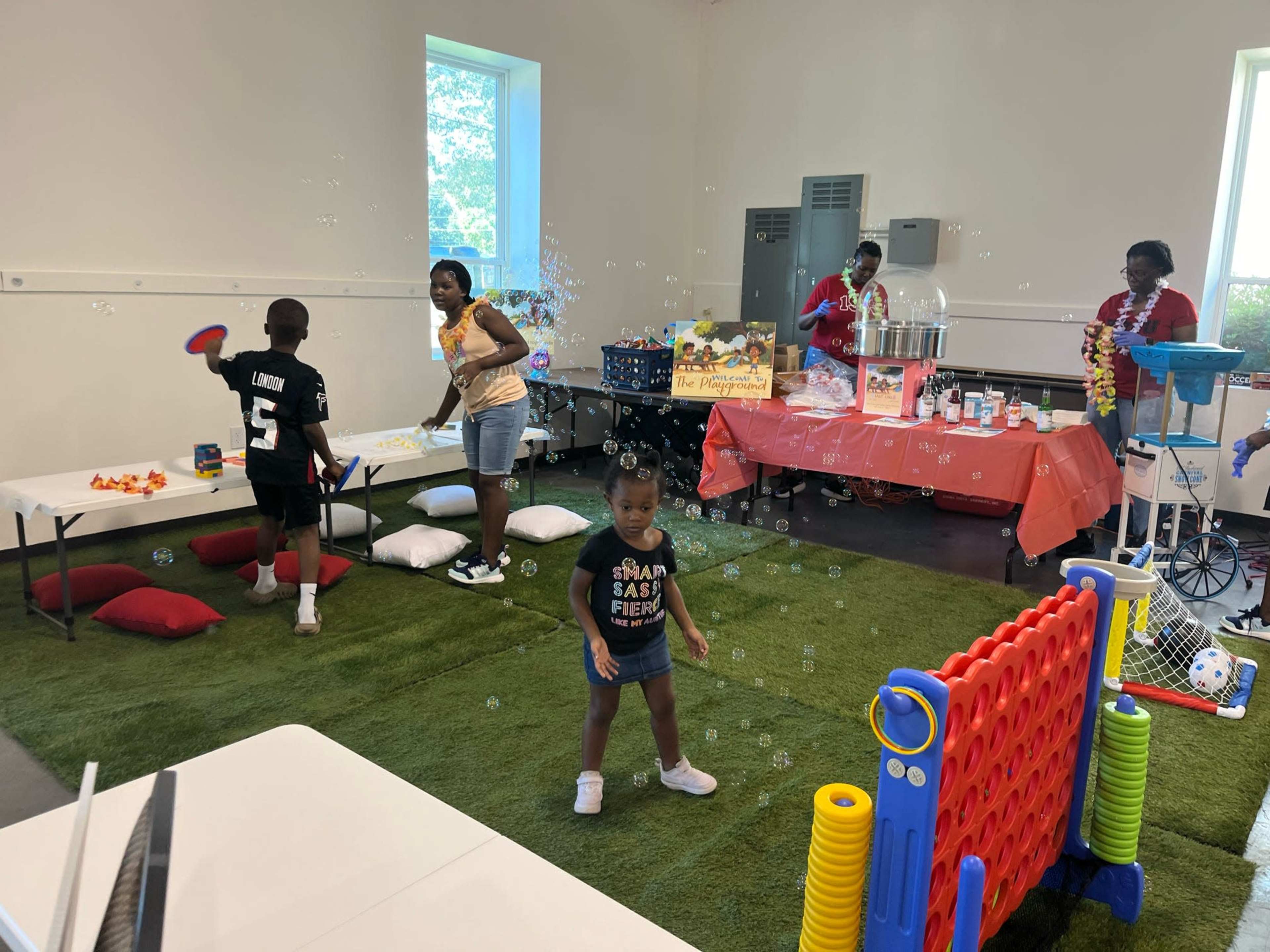Children are playing with bubbles in a colorful activity room, while adults prepare snacks and decorations in the background.
