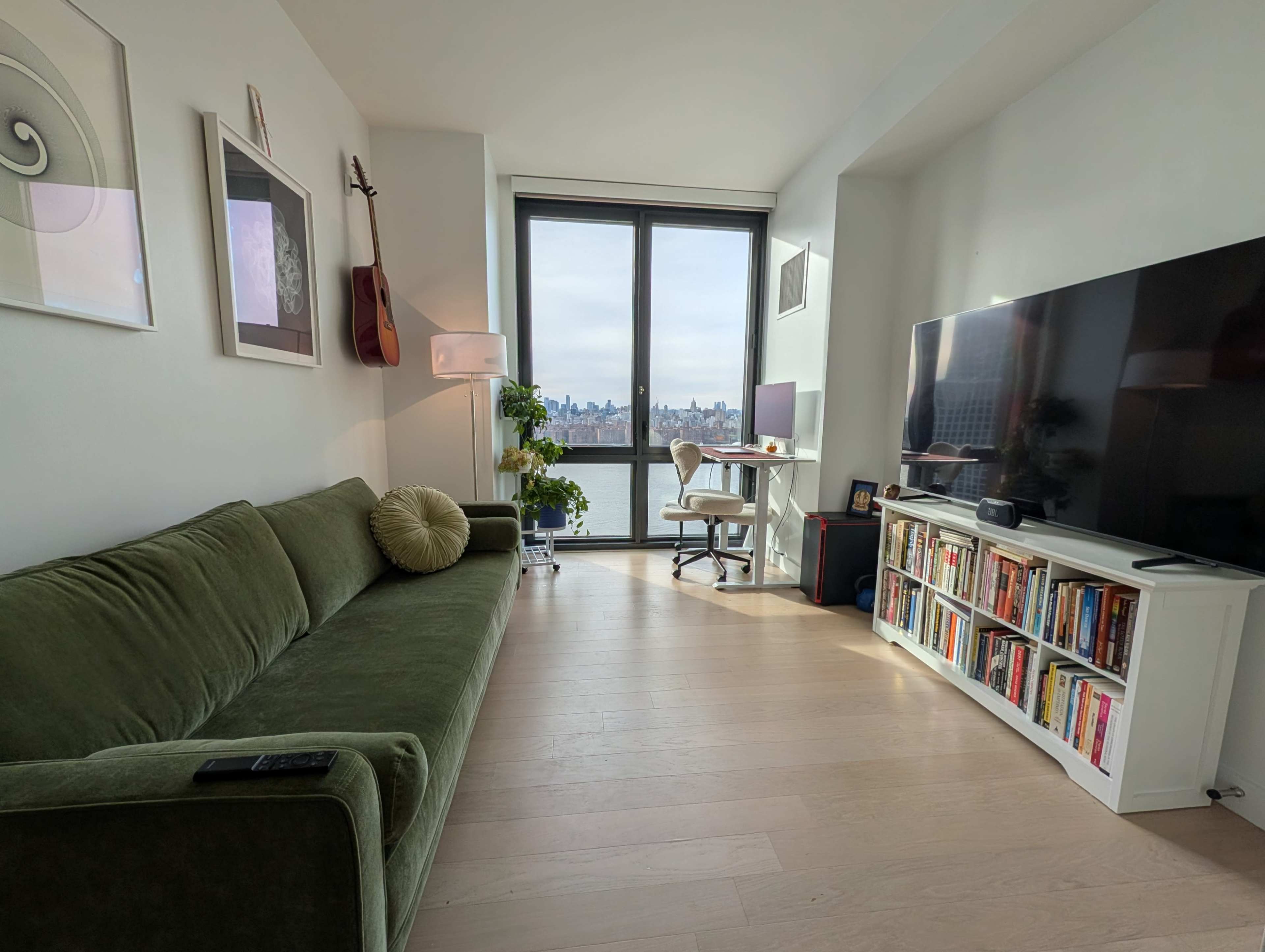 The room features a green sofa, a lamp, a television on a white stand, and a desk, all arranged near large windows overlooking a view of the city.