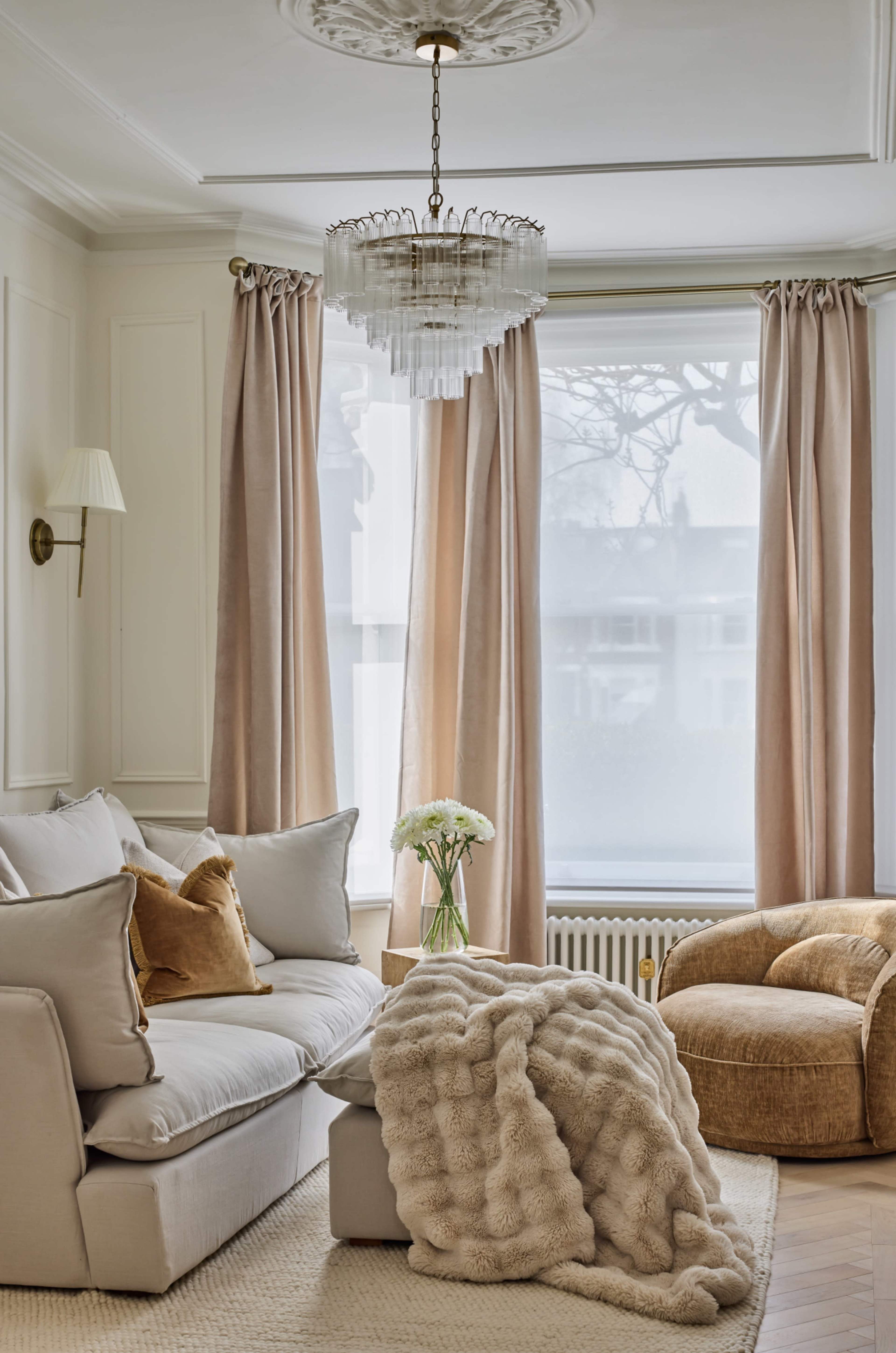 The image depicts a stylish living room with a light-colored sofa, a velvet armchair, and large windows dressed with soft curtains, accented by a textured throw blanket and a floral arrangement on a coffee table.