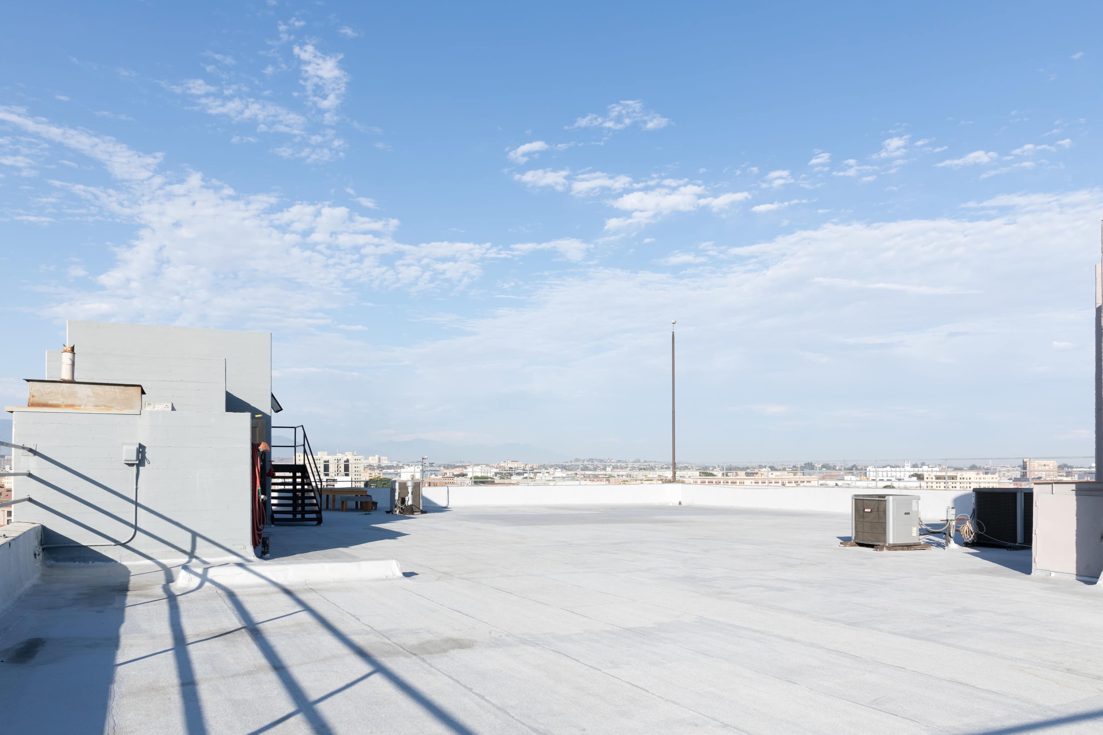 The image shows a flat rooftop with air conditioning units, a staircase leading down, and a clear blue sky in the background.