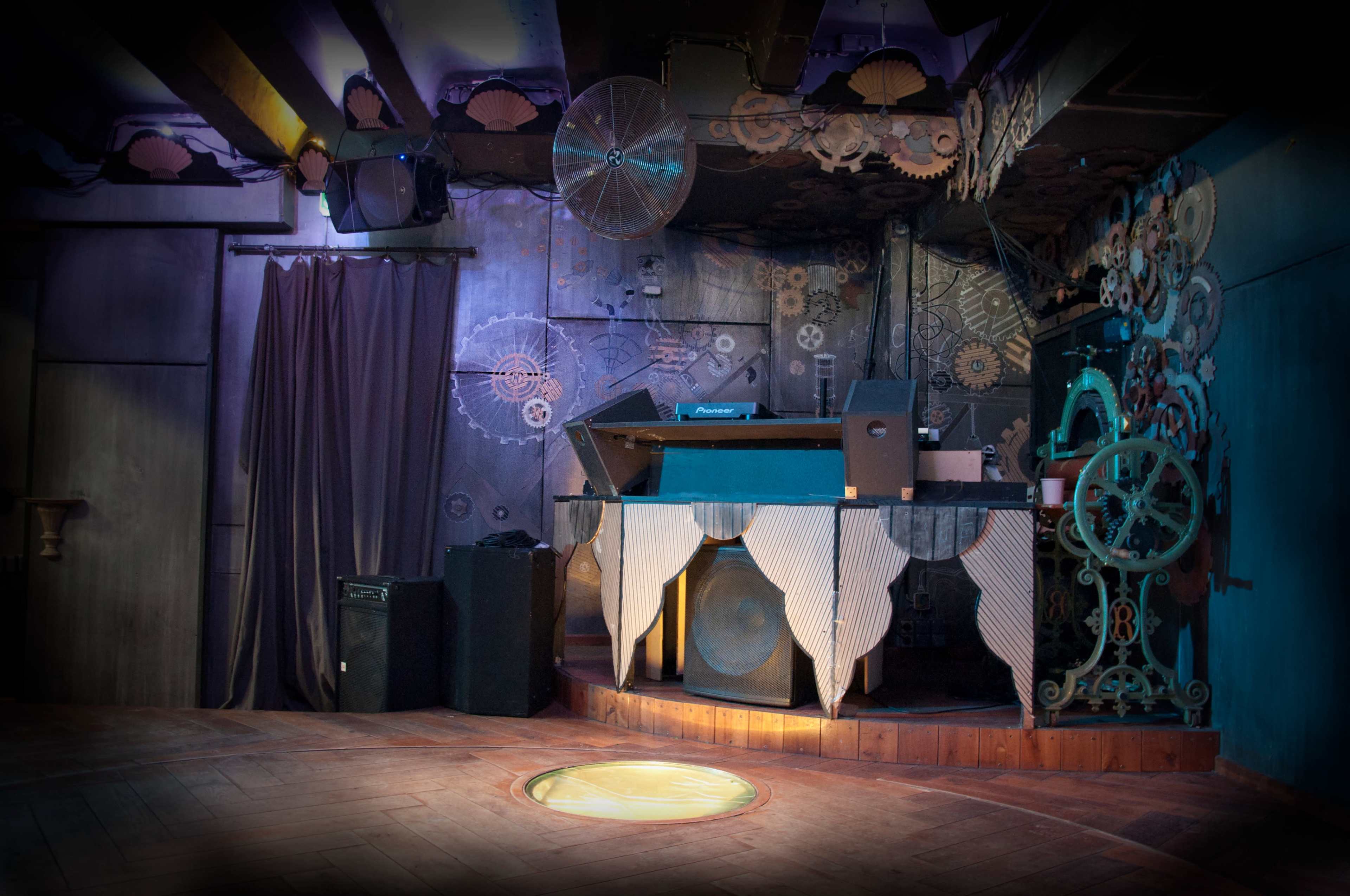 The image shows a dimly lit DJ booth decorated with mechanical gears and surrounded by a wooden floor in a club setting.