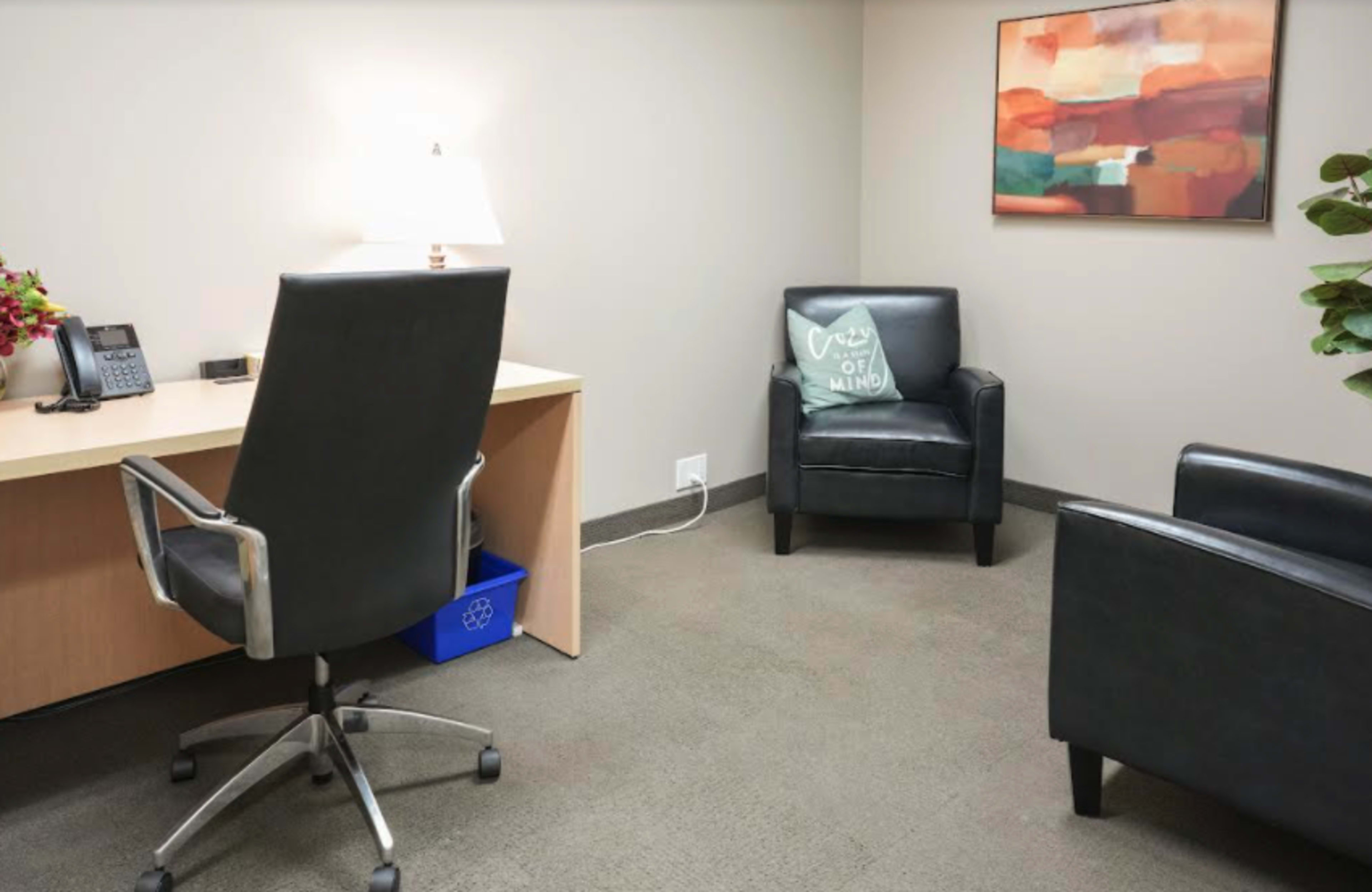 The image shows a small office space with a desk and chair, a lamp on the desk, two armchairs, and a piece of wall art.