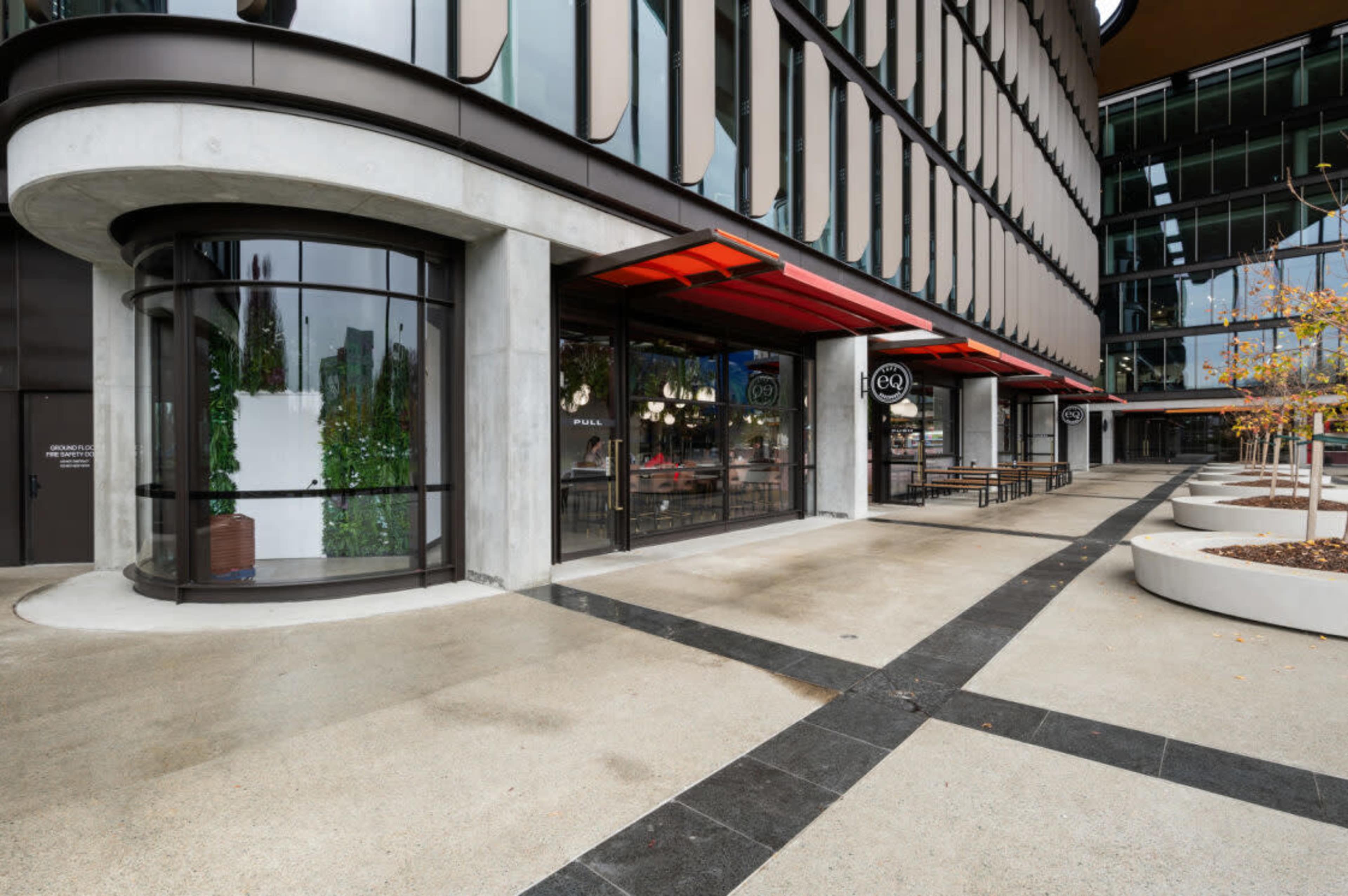 The image shows a modern building with large glass windows and a paved walkway, featuring seating areas outside and decorative plants along the entrance.