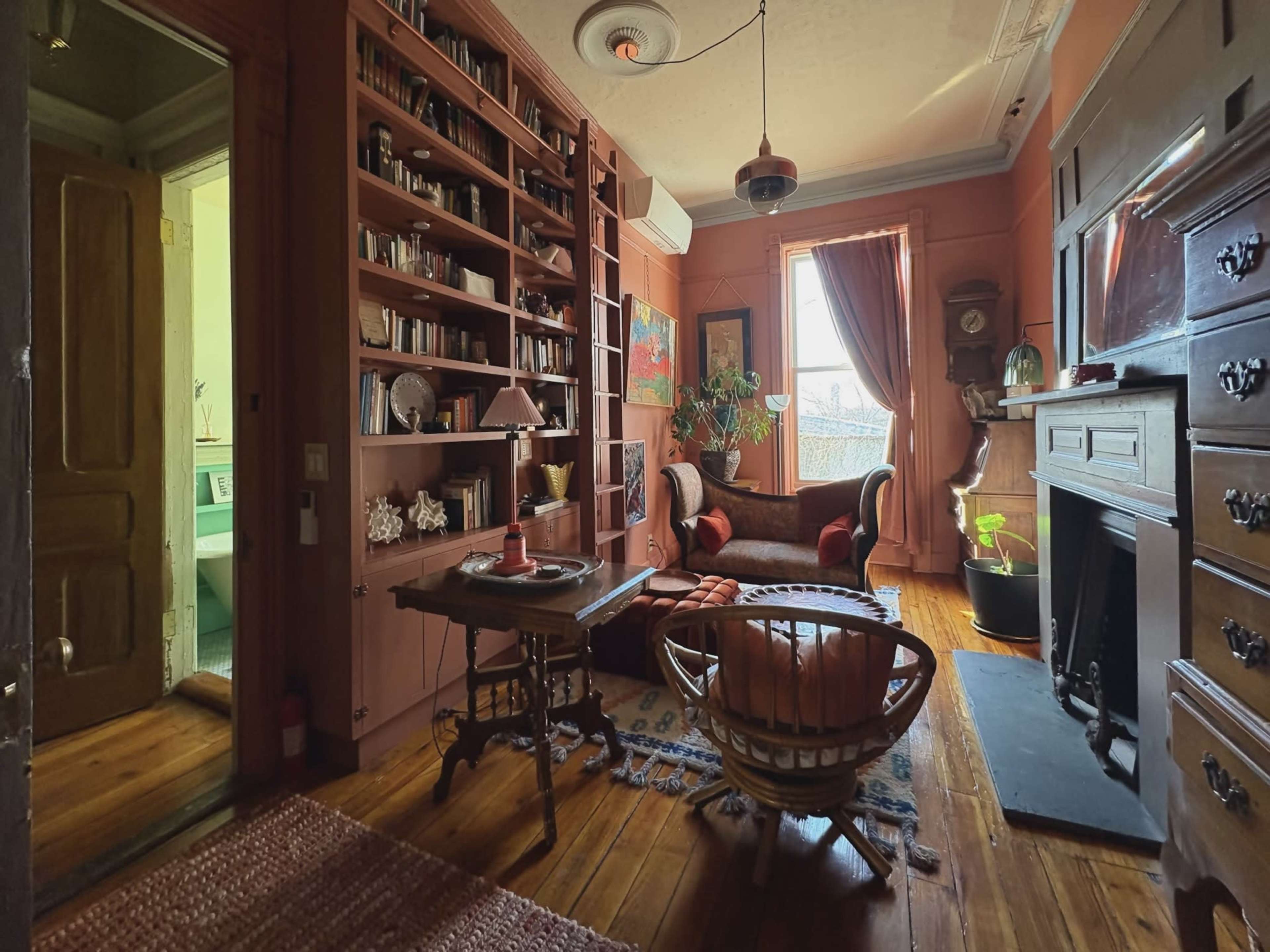 The image shows a cozy sitting room with bookshelves, a vintage couch, and a decorative fireplace, set in warm colors and featuring wooden floors.