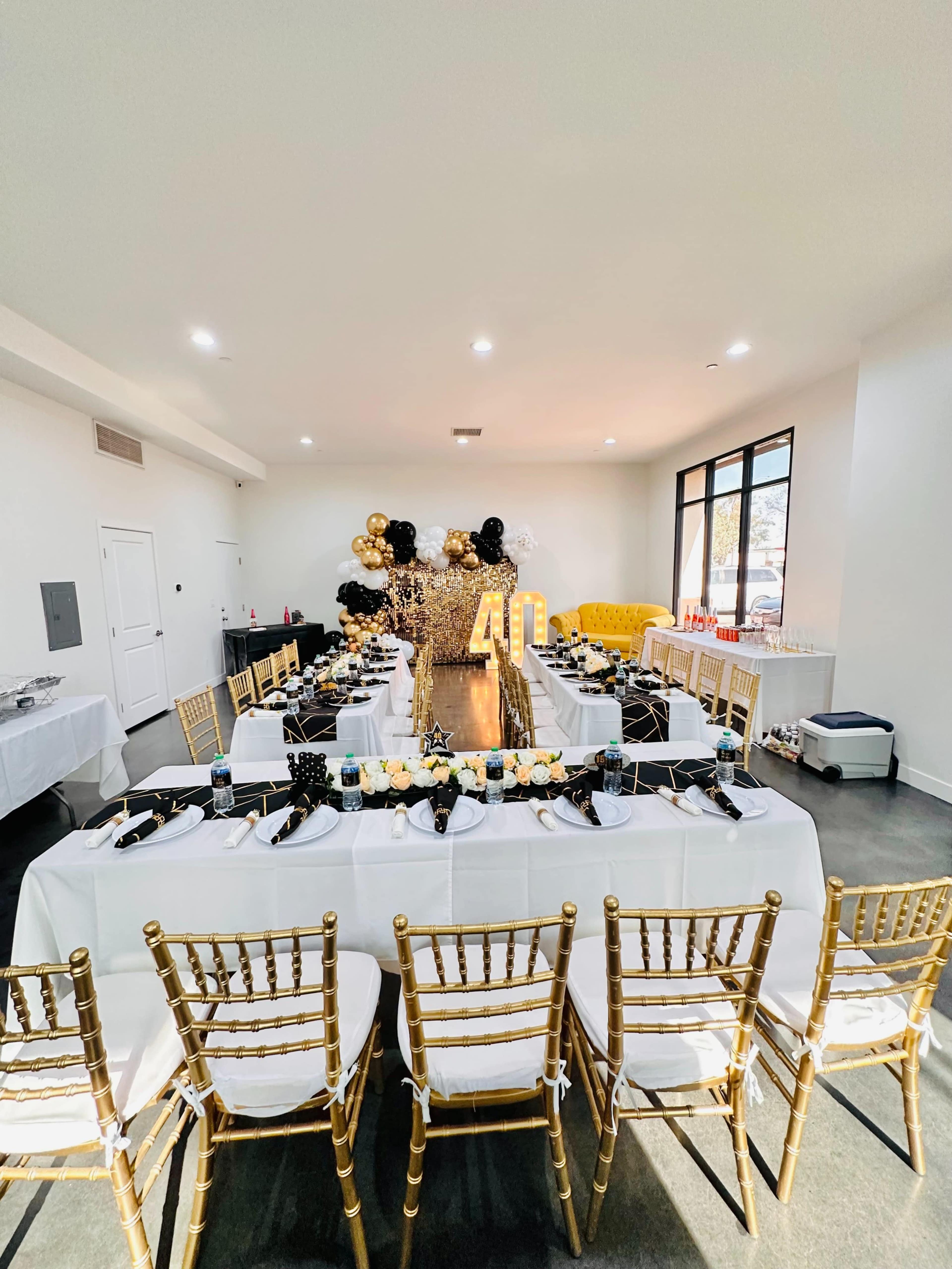 A dining area is set up with several long tables draped in white linens, adorned with black and gold decorations, and surrounded by gold Chiavari chairs.