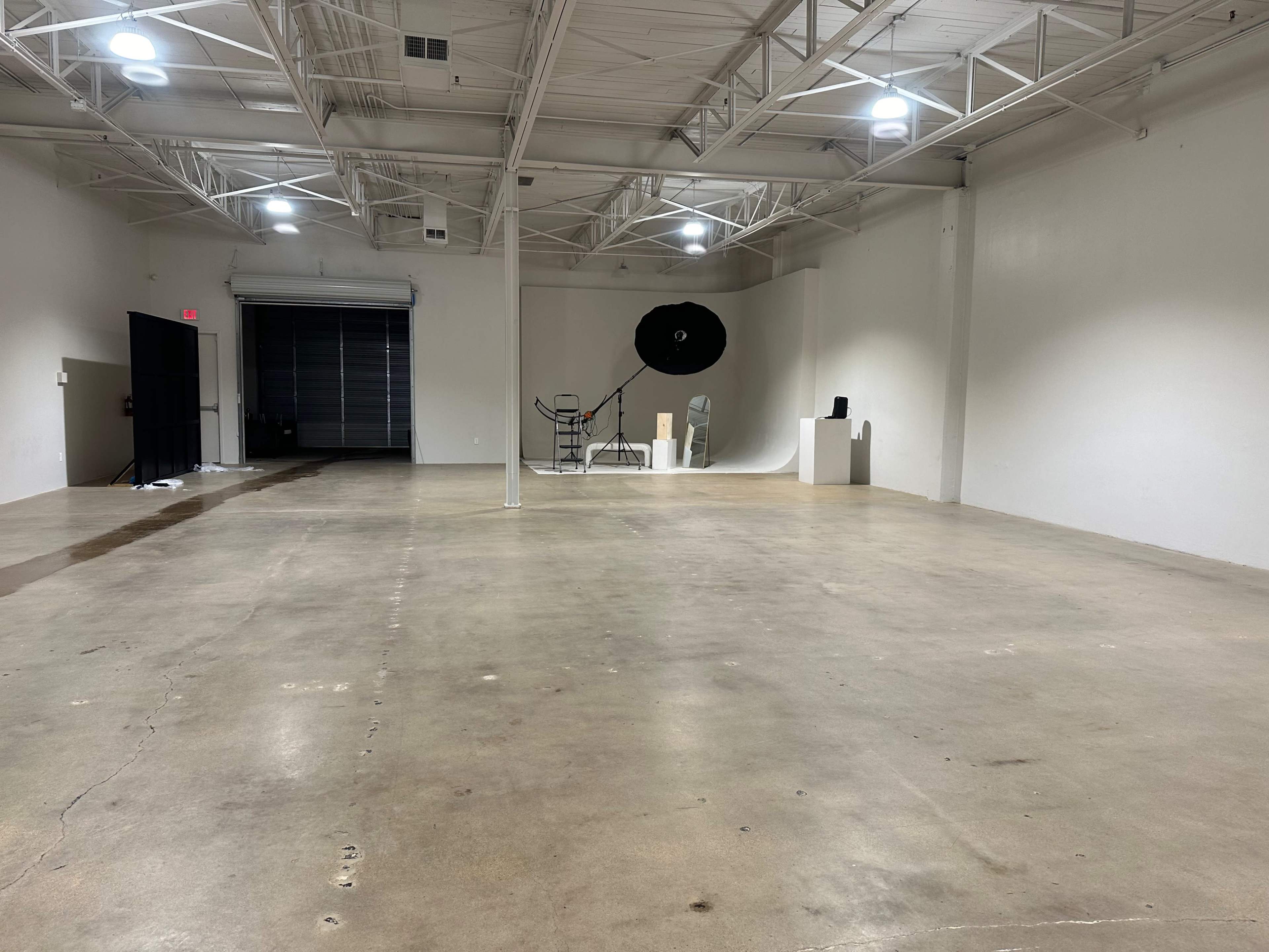 A spacious, empty commercial interior with a polished concrete floor and minimal furnishings, including a photography backdrop and equipment setup against the far wall.