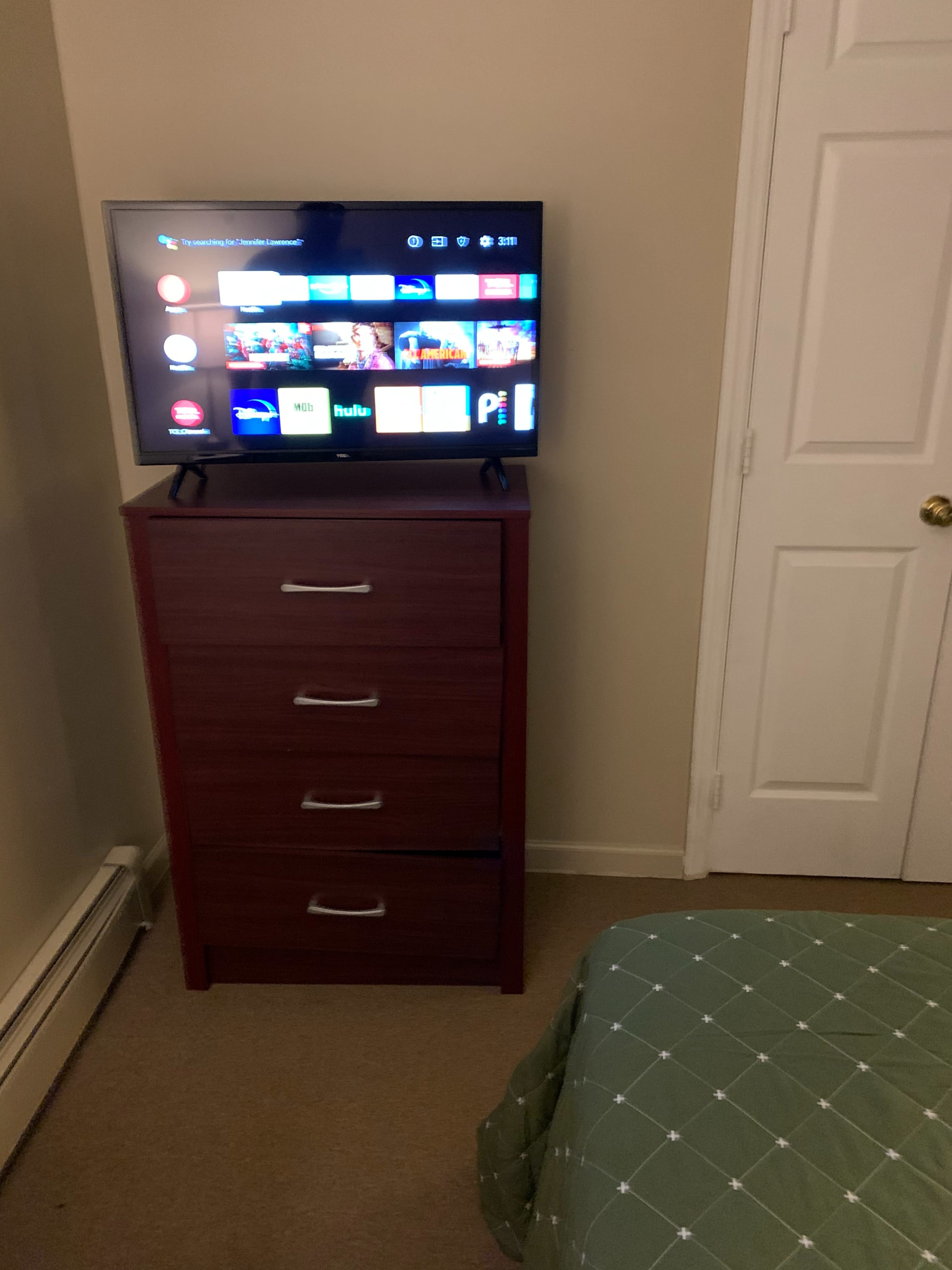 A television is mounted on a dresser next to a closed door in a small bedroom with a bed covered by a green patterned blanket.