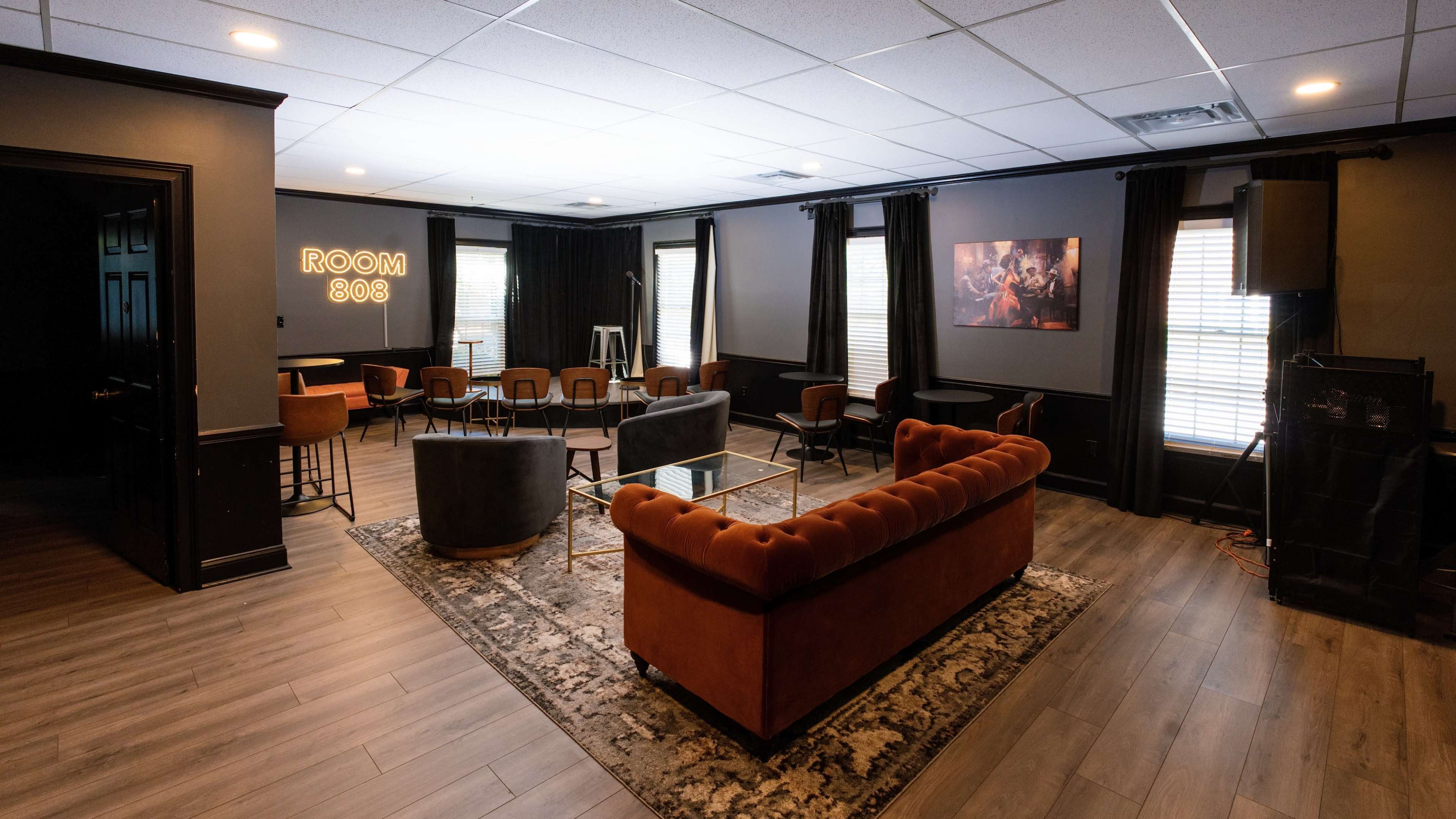 The image shows a lounge area with a modern design featuring a brown velvet sofa, seating arrangements, and large windows with dark curtains.