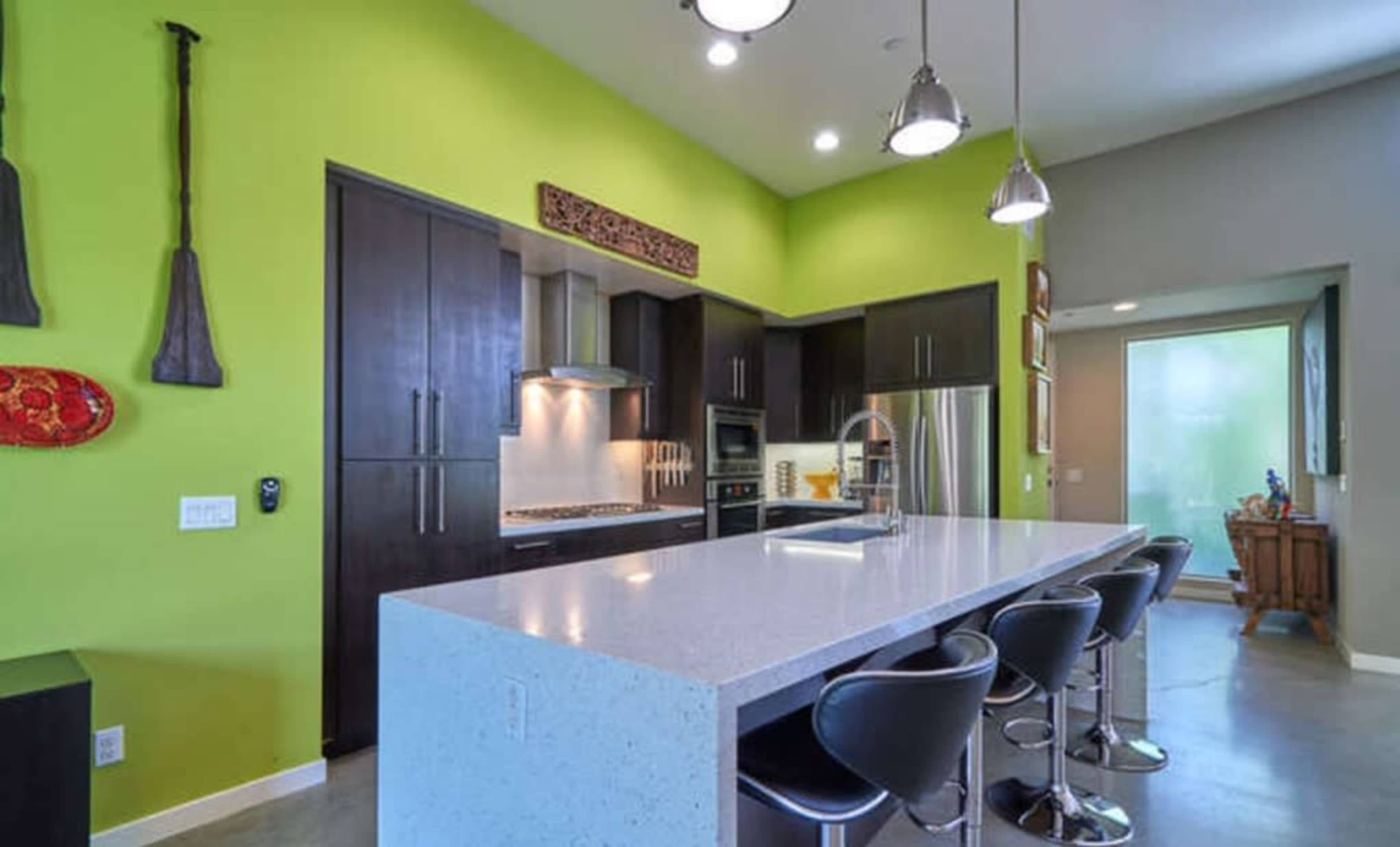 A modern kitchen featuring a green accent wall, a large white countertop island with black bar stools, and stainless steel appliances.