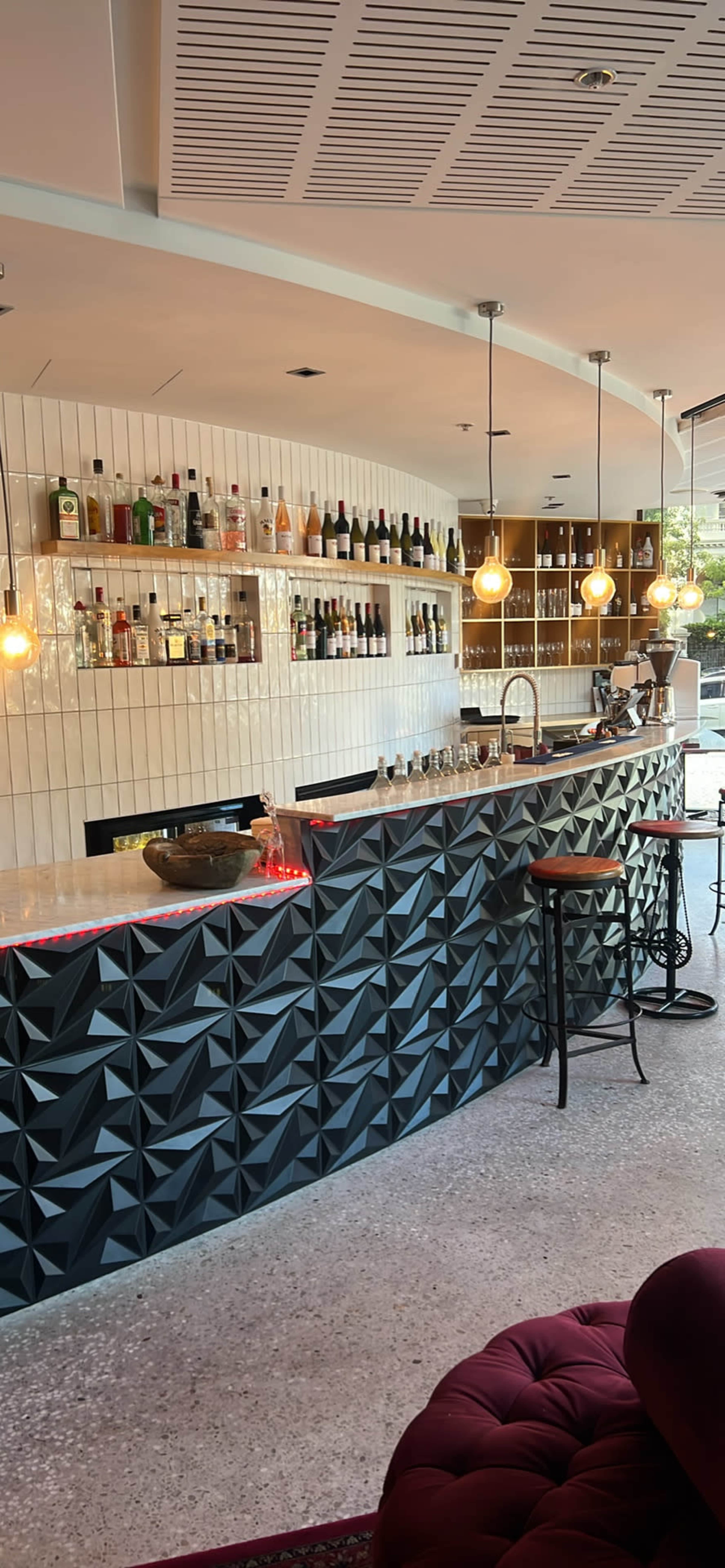 The image shows a modern bar area with a geometric patterned counter, bottles of various liquors displayed on shelves, and pendant lights hanging above.