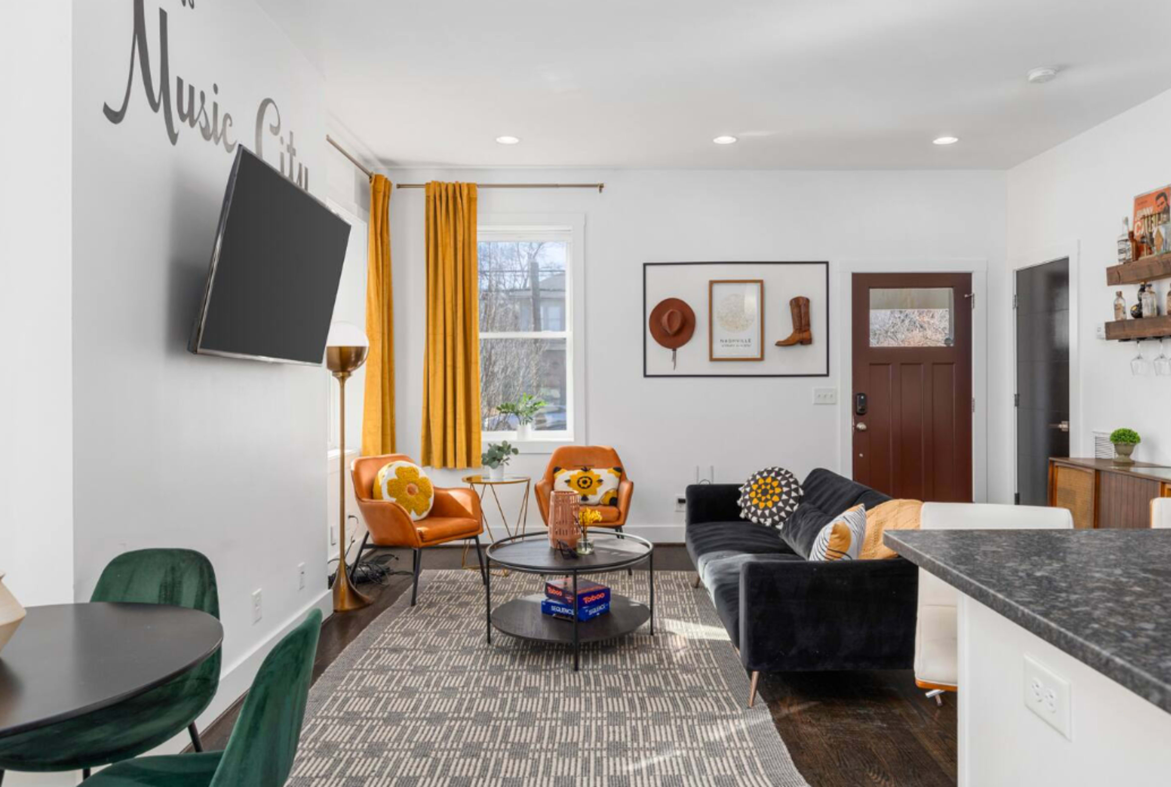 A modern living room with a black sofa, orange armchairs, a round coffee table, and large windows allowing natural light, accented by vibrant decor and a TV mounted on the wall.
