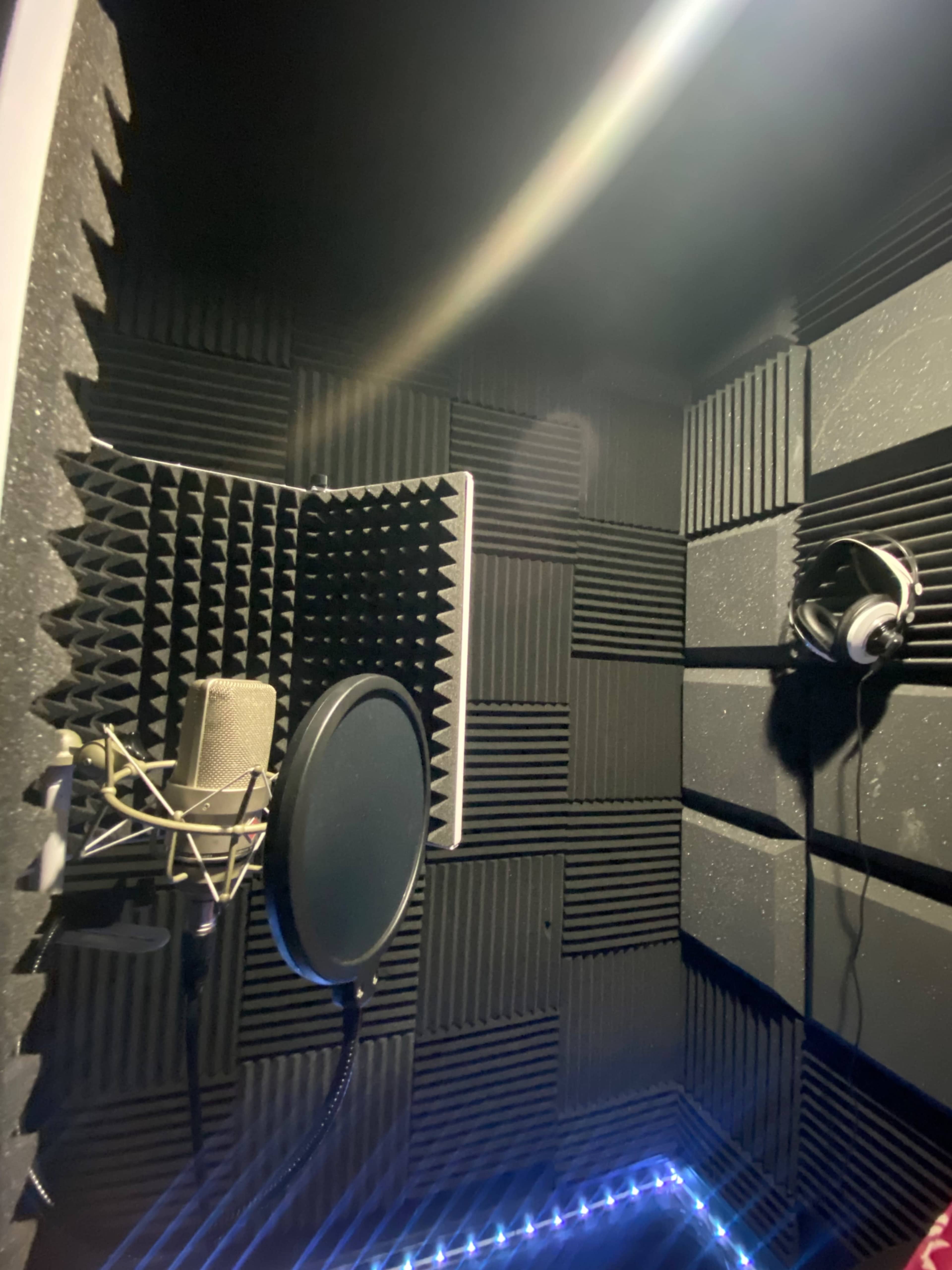 A soundproof recording studio booth featuring acoustic foam panels, a microphone on a stand, and a pair of headphones hung on the wall.