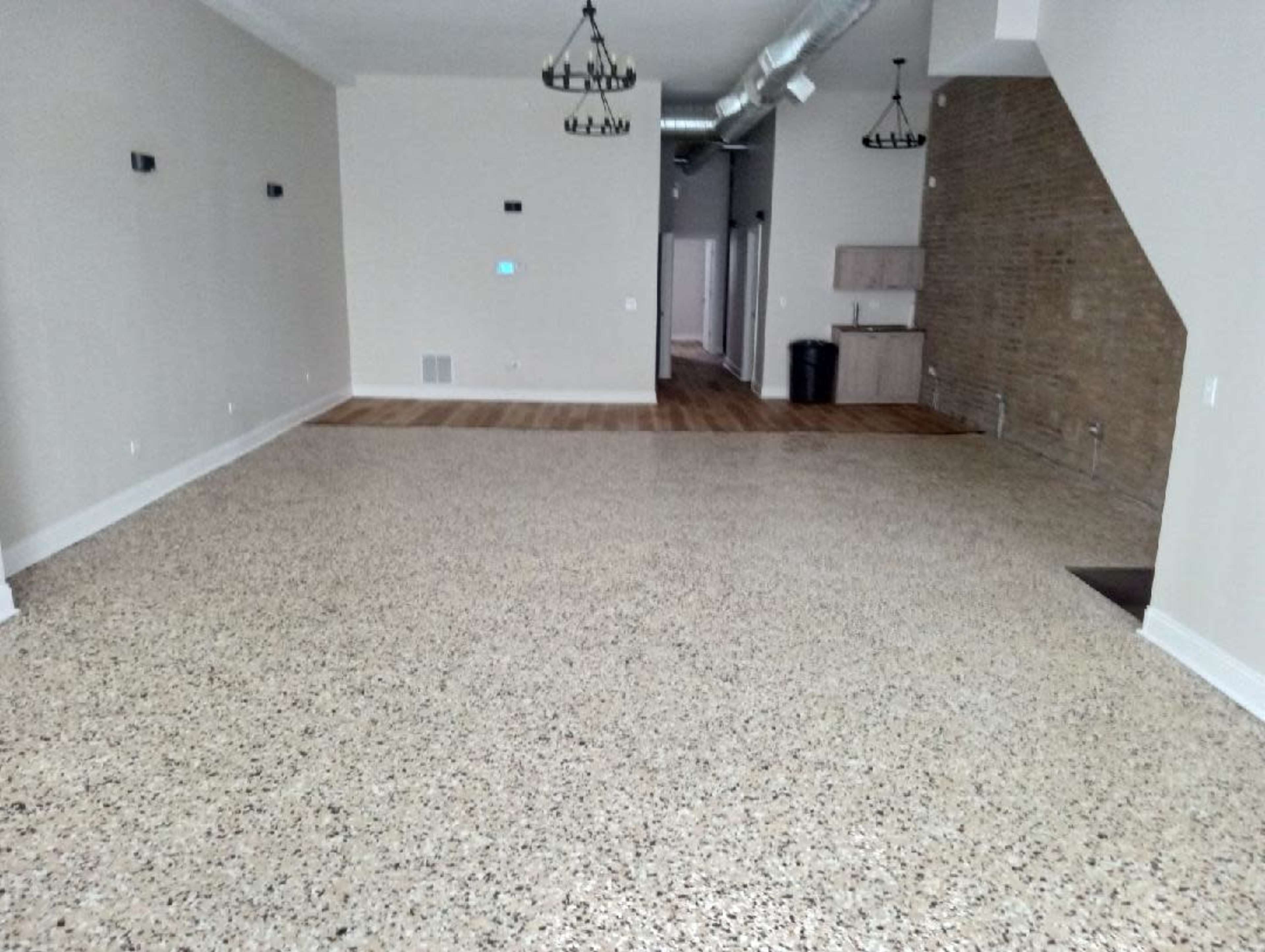 The image shows a spacious, empty room with polished concrete flooring, a light-colored wall, and an adjoining kitchen area in the background.