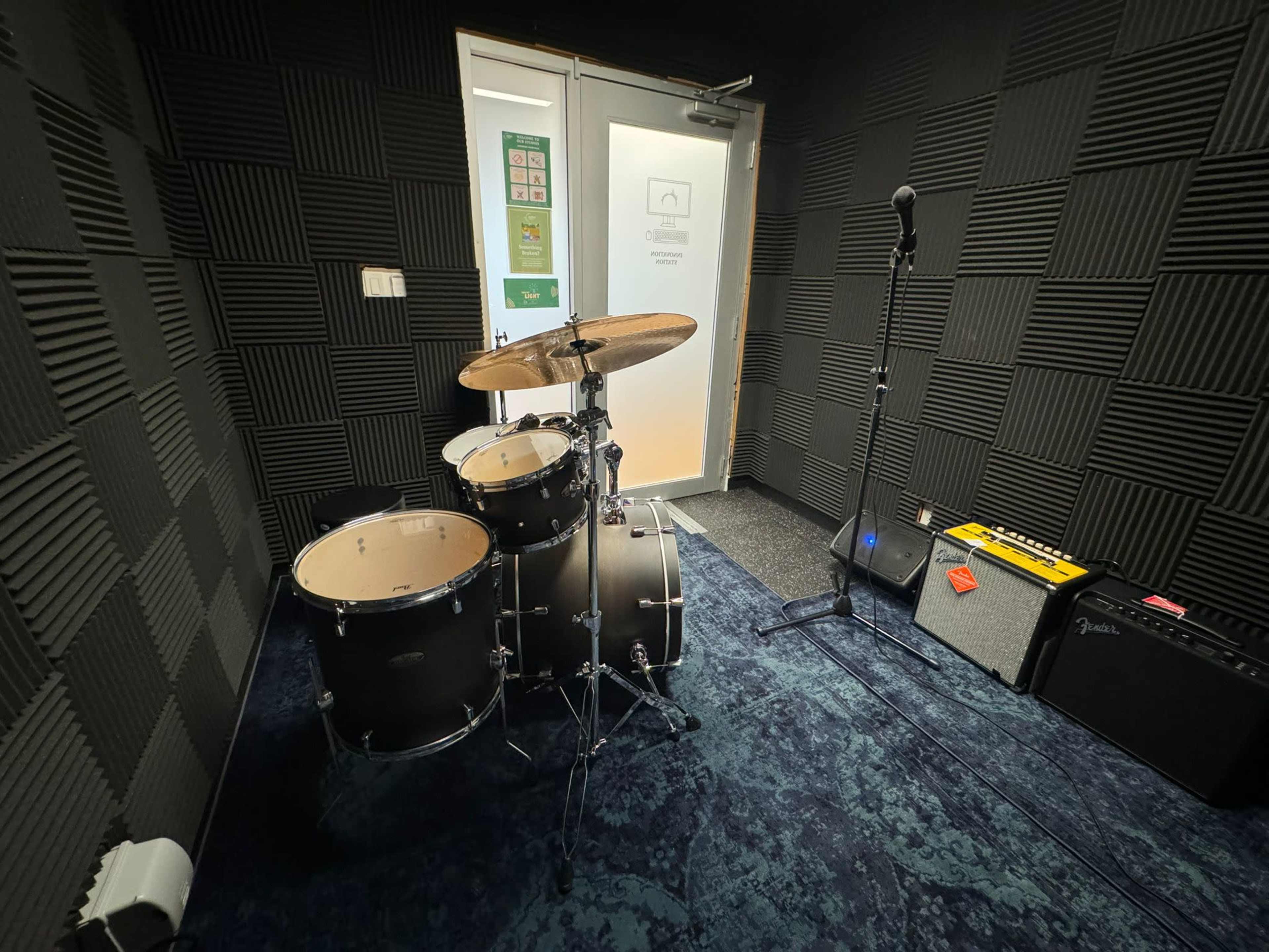 A soundproofed music practice room featuring a drum set, microphone stand, amplifier, and a carpeted floor.