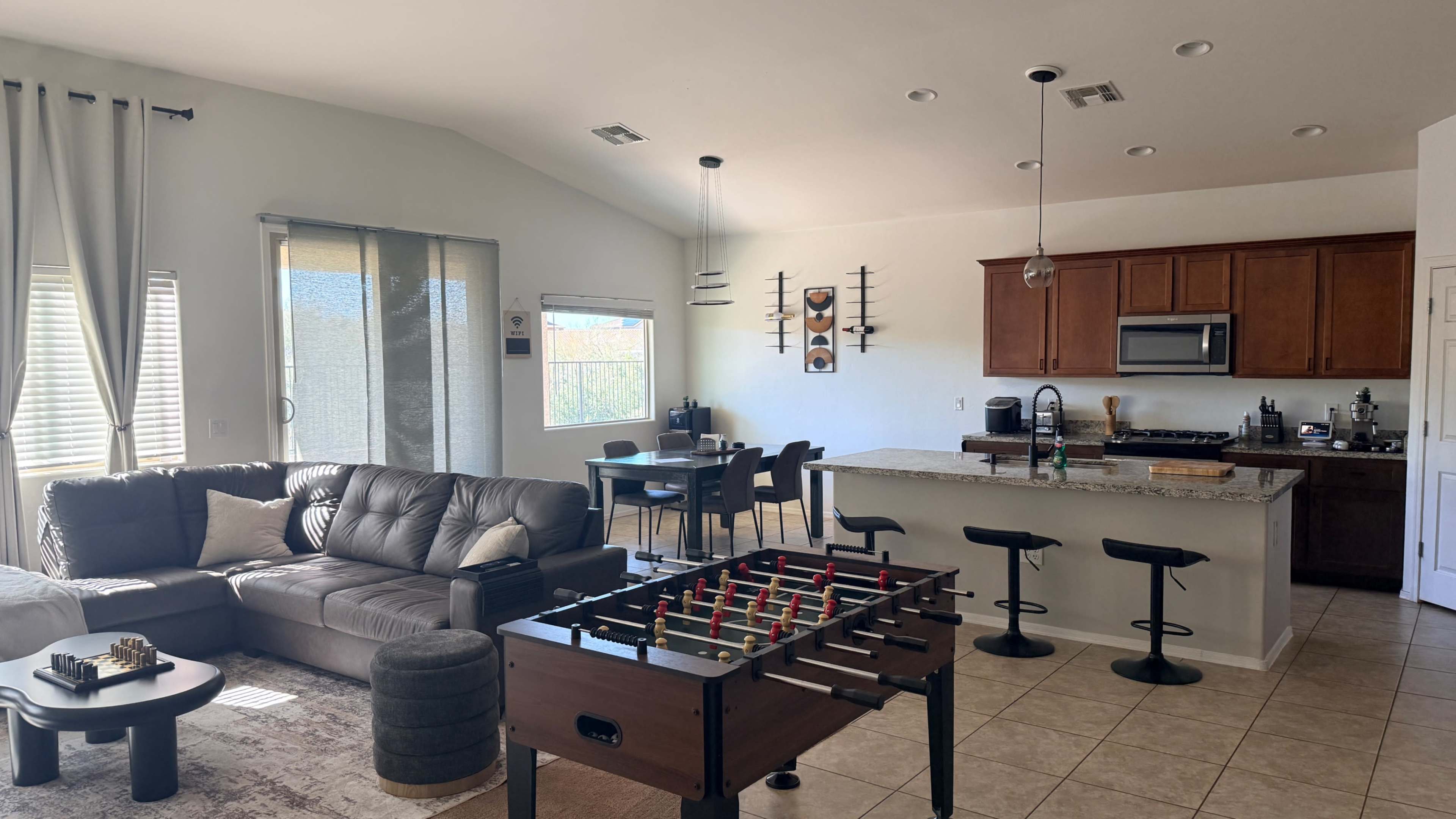 The image shows a modern living space featuring a leather sectional sofa, a foosball table, and an open kitchen with wooden cabinets and a dining area.