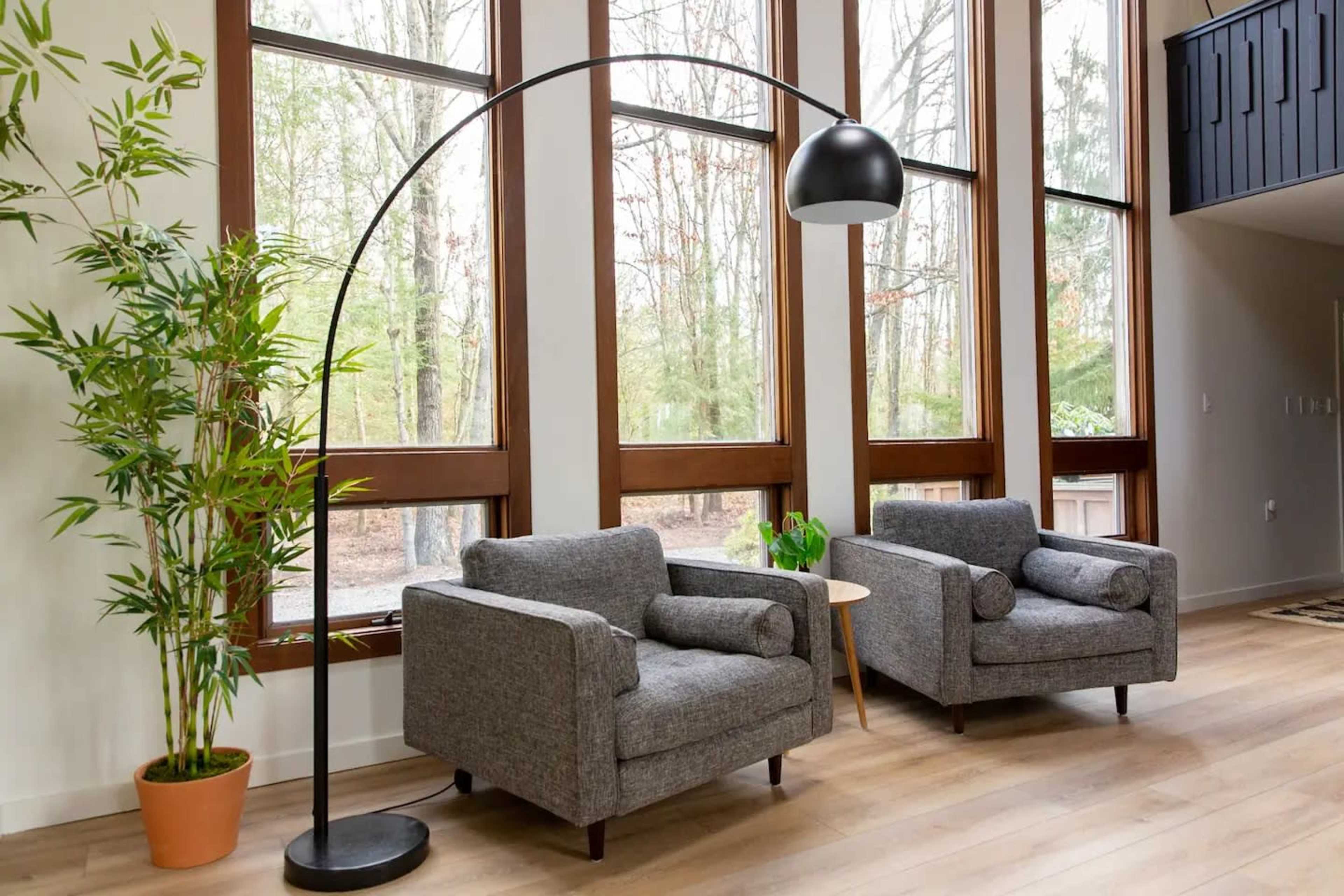 Two gray armchairs with round cushions, positioned beside a curved floor lamp and a small table, set against large windows overlooking a forested area.
