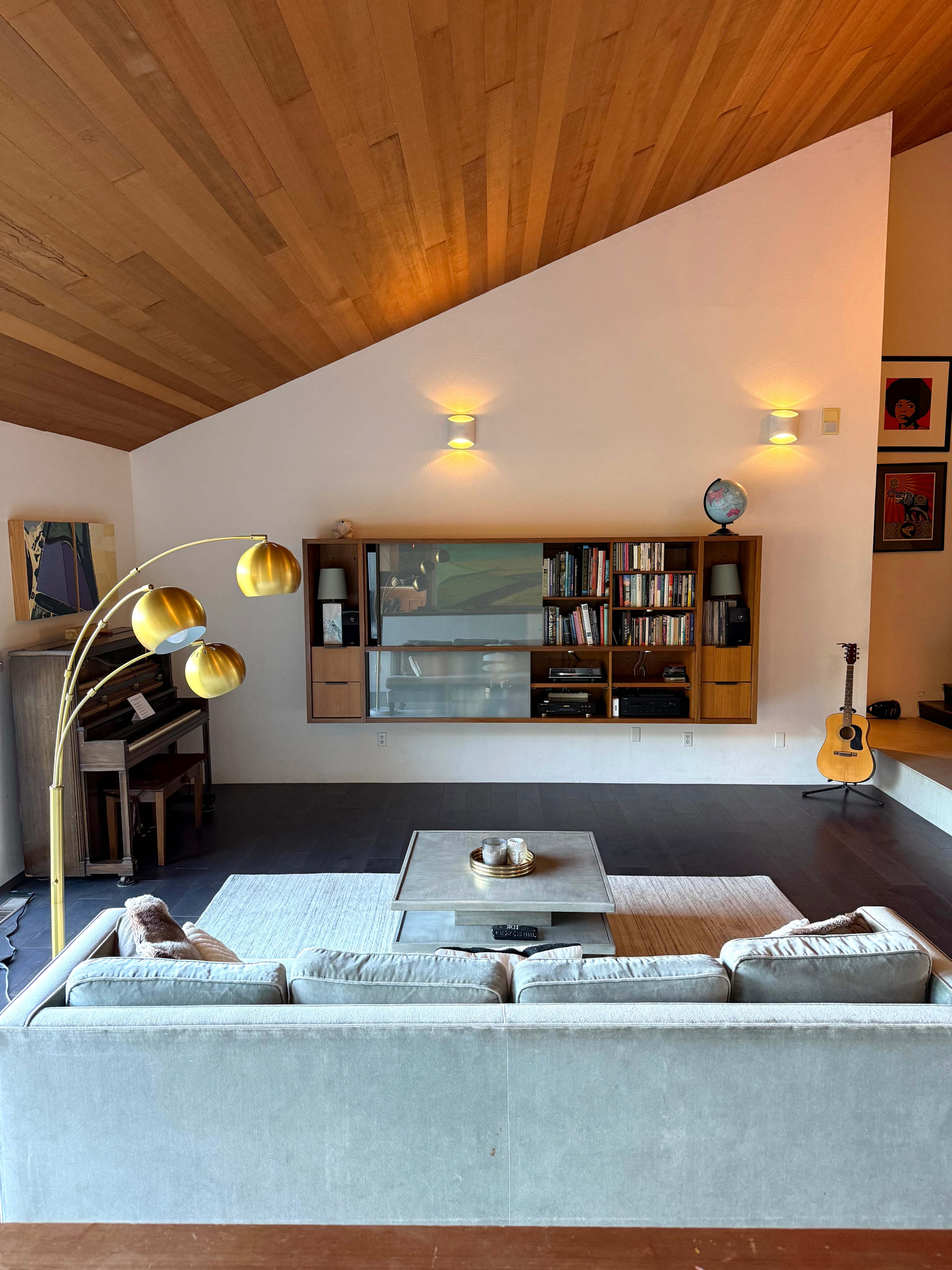The image shows a modern living room featuring a light gray sofa, a coffee table, a wall-mounted bookshelf, and a guitar leaning against the wall.