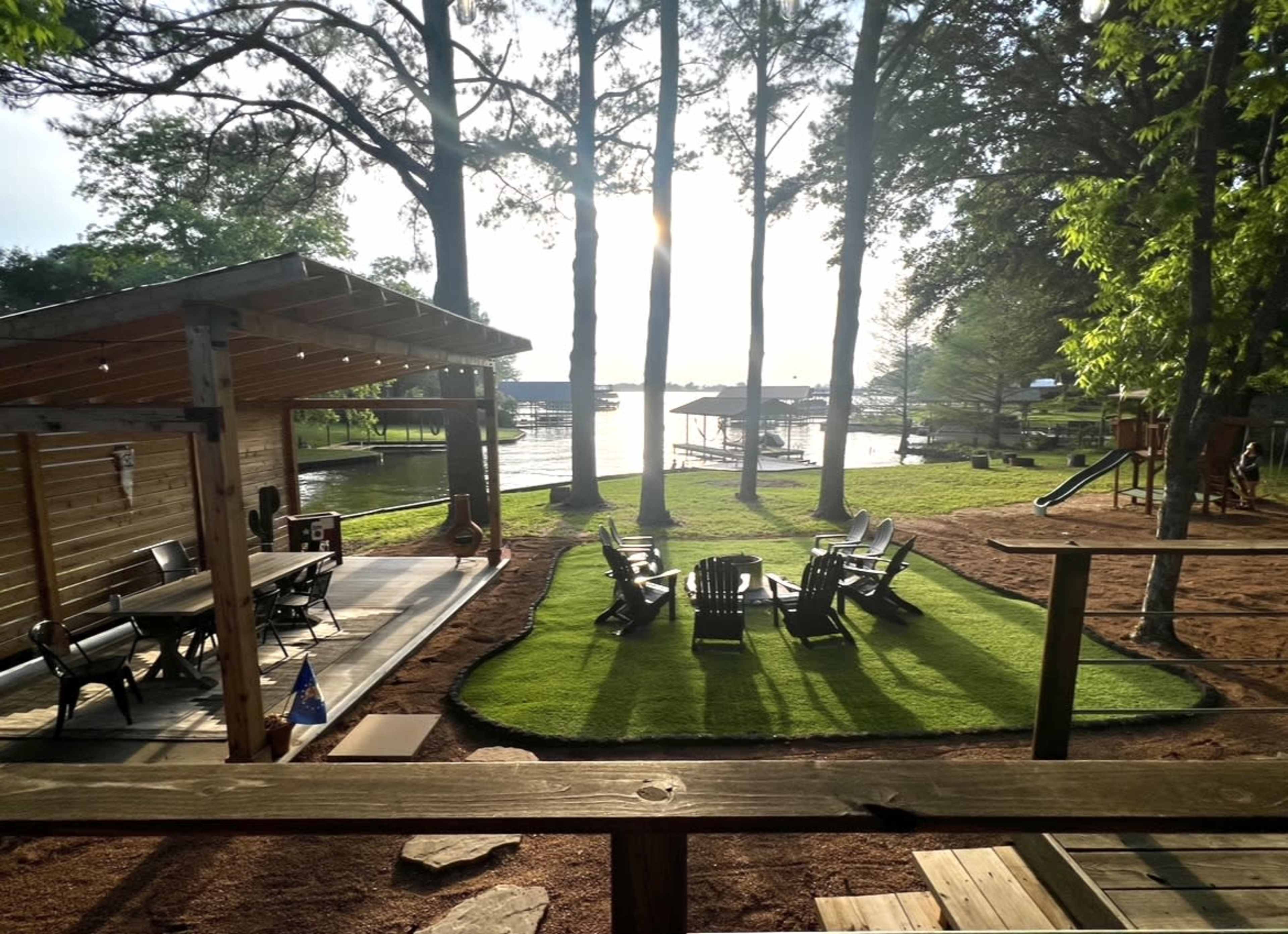 A lakeside area with a grassy space surrounded by trees, featuring a seating arrangement of adirondack chairs and a dock in the background.
