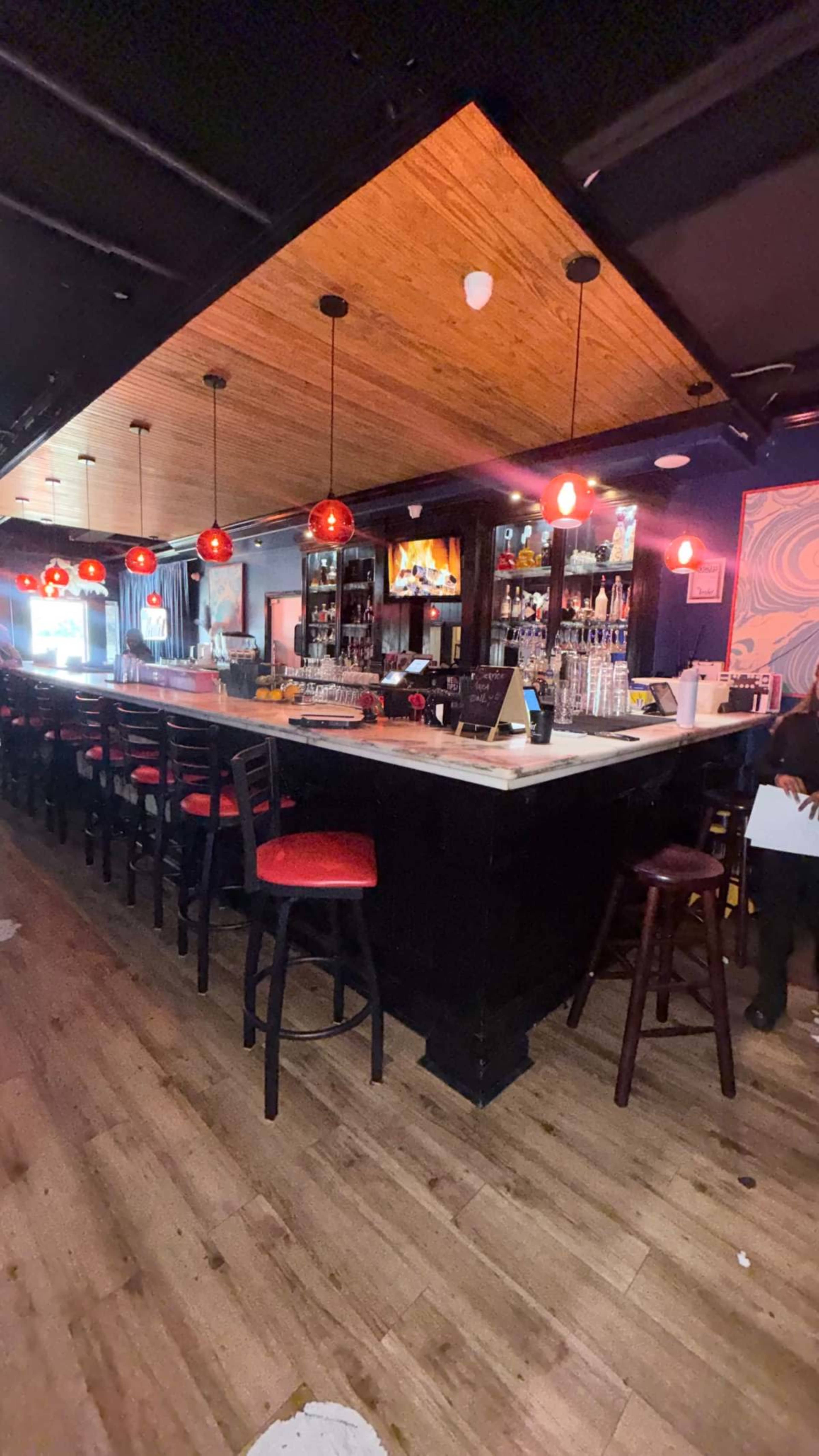 The image shows the interior of a bar with a long counter, red pendant lights, and high stools aligned along the bar.