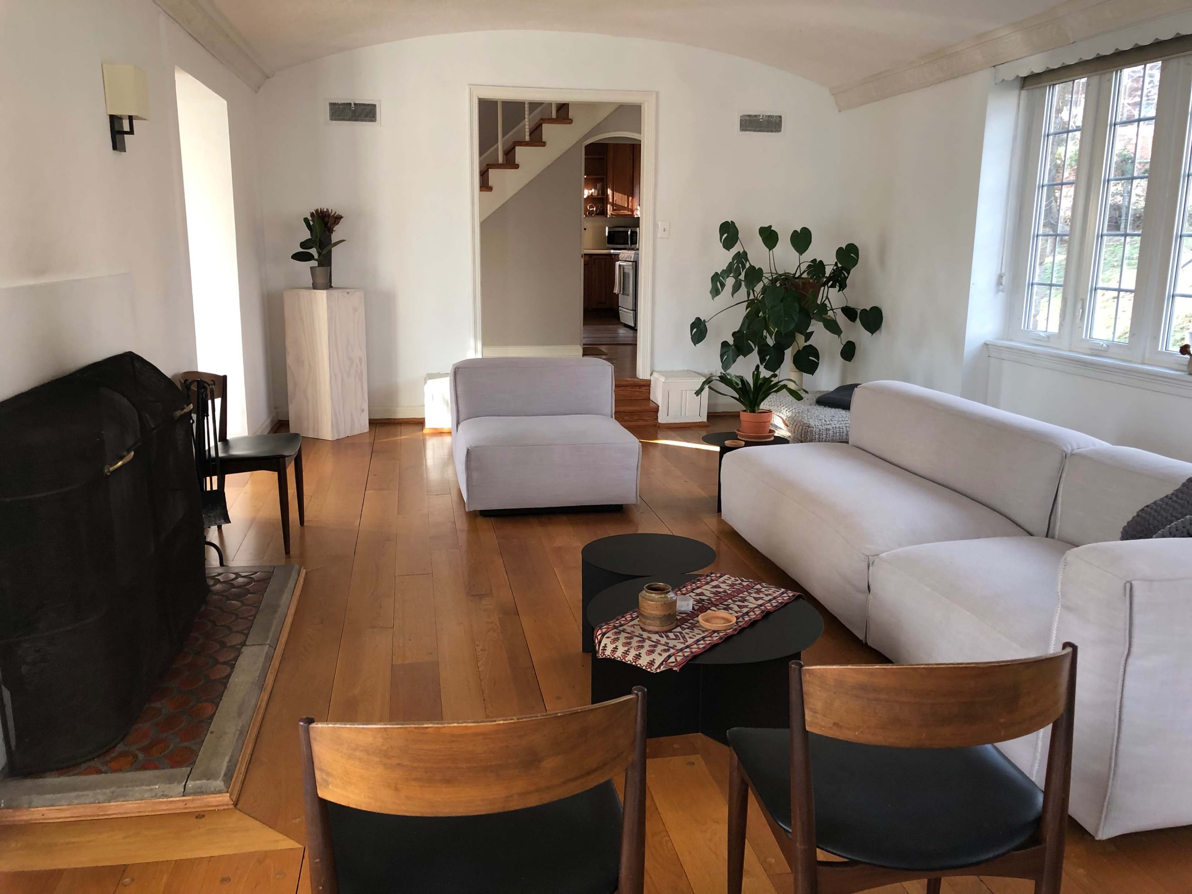 A bright living room with a fireplace, a gray sofa, two armchairs, a coffee table, and a view of a staircase leading to the kitchen.
