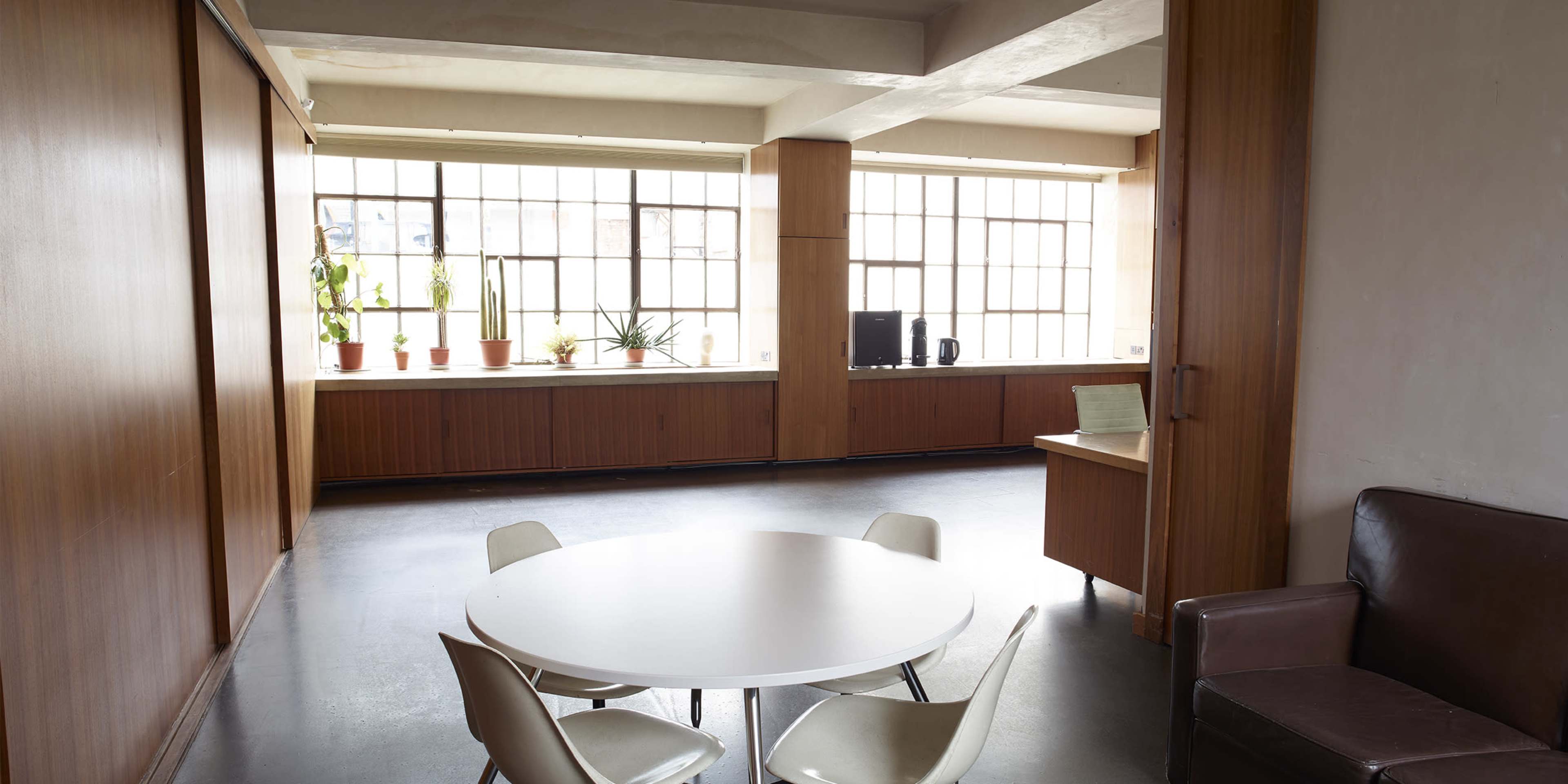 The image shows a spacious interior office area with a round table, four chairs, a brown couch, potted plants on the windowsill, and a desk with a computer.