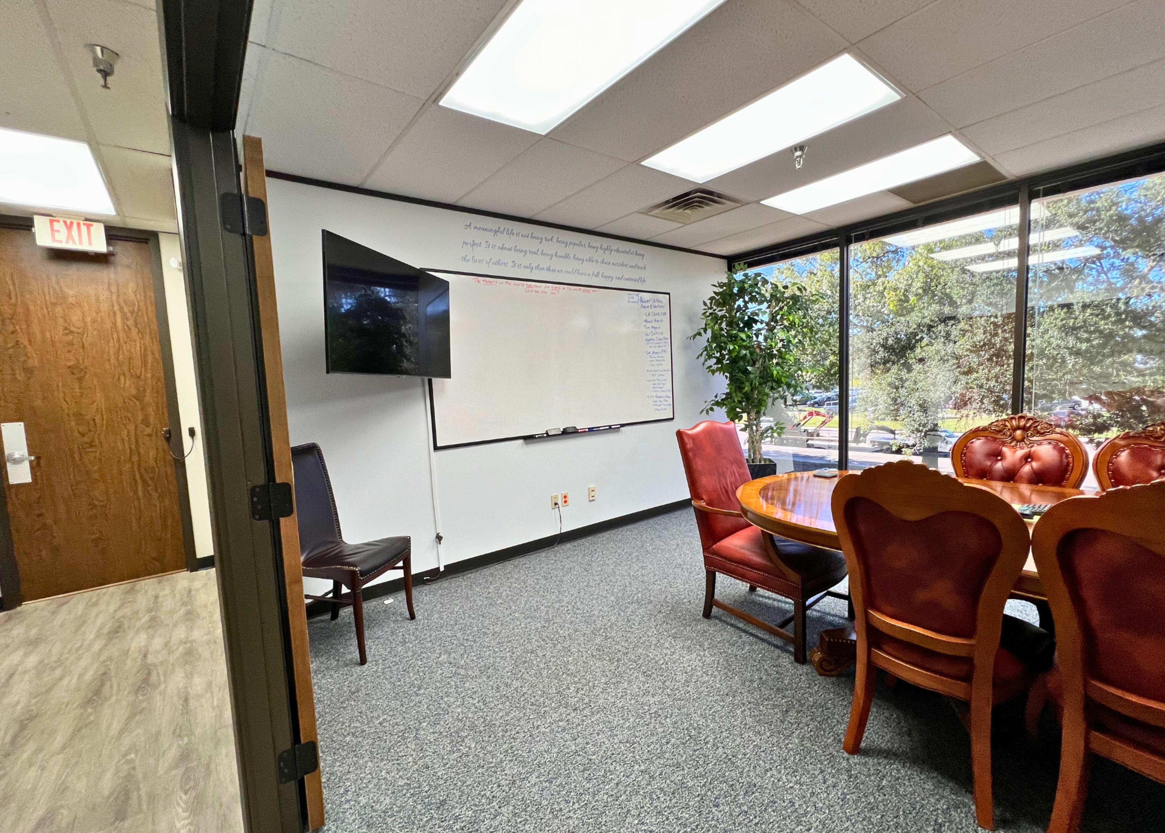A conference room features a round table with high-backed chairs, a whiteboard on the wall, and a television mounted nearby, illuminated by natural light from large windows.