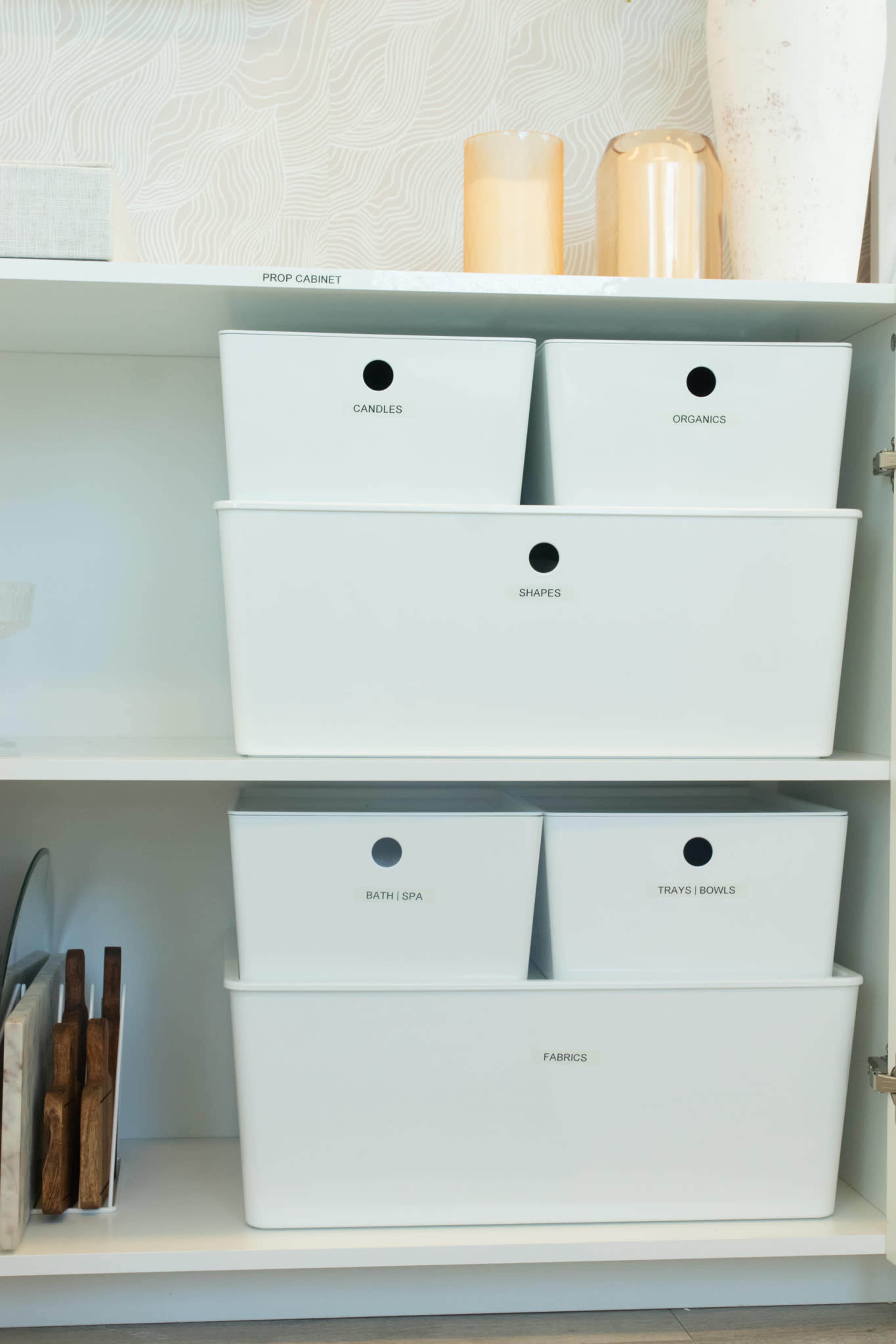 A neatly organized cabinet featuring labeled white storage bins containing various items such as candles, organic materials, and fabric.